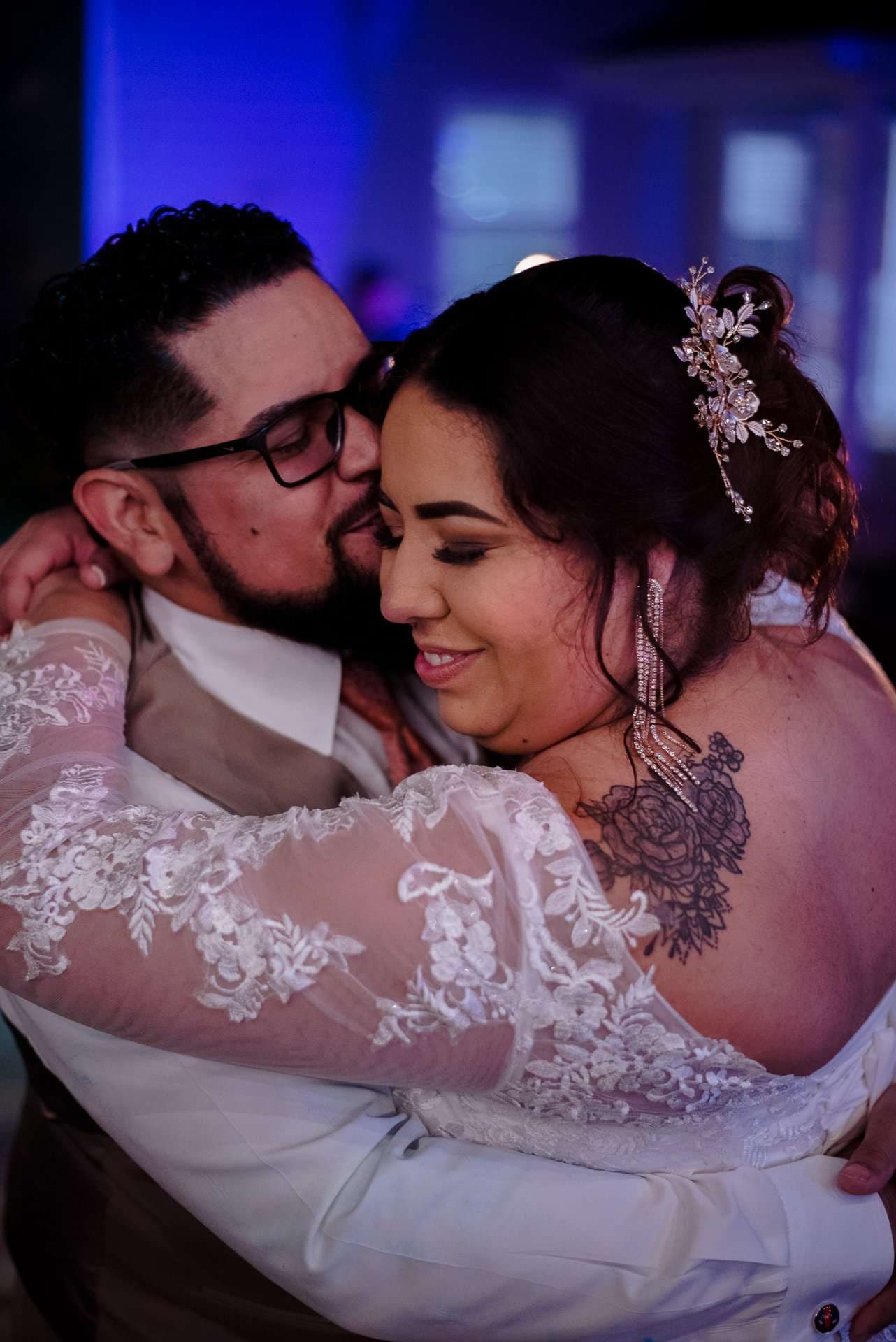Bride in a white lace gown with a floral back tattoo embraces a bearded groom in glasses under blue lighting