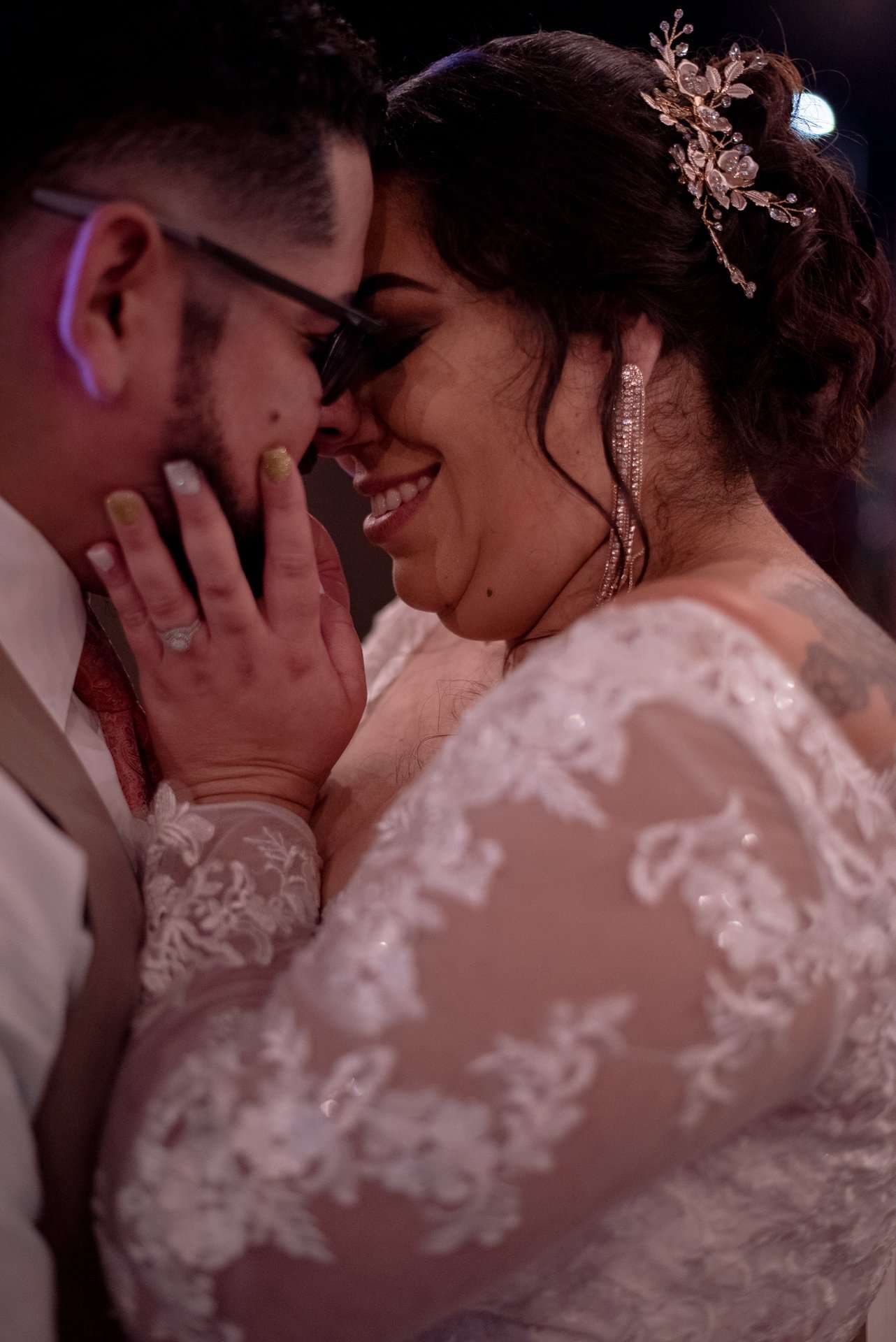 Smiling bride in a lace gown and floral hair piece touches foreheads with her partner, her hand on his cheek