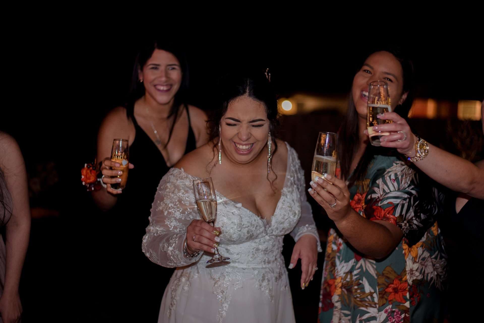 Laughing bride in a white lace gown holding a champagne flute, surrounded by smiling guests raising drinks at night