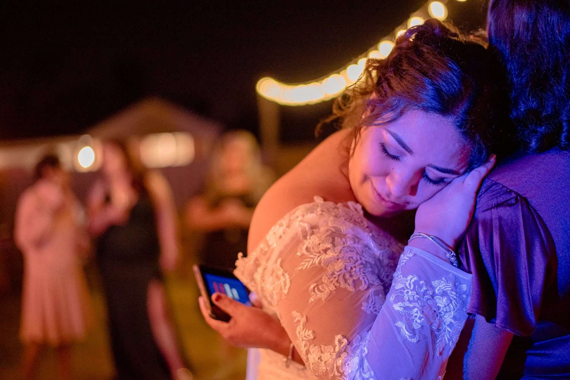 Bride in a white lace gown rests her head on a guest's shoulder, eyes closed, holding a phone under string lights