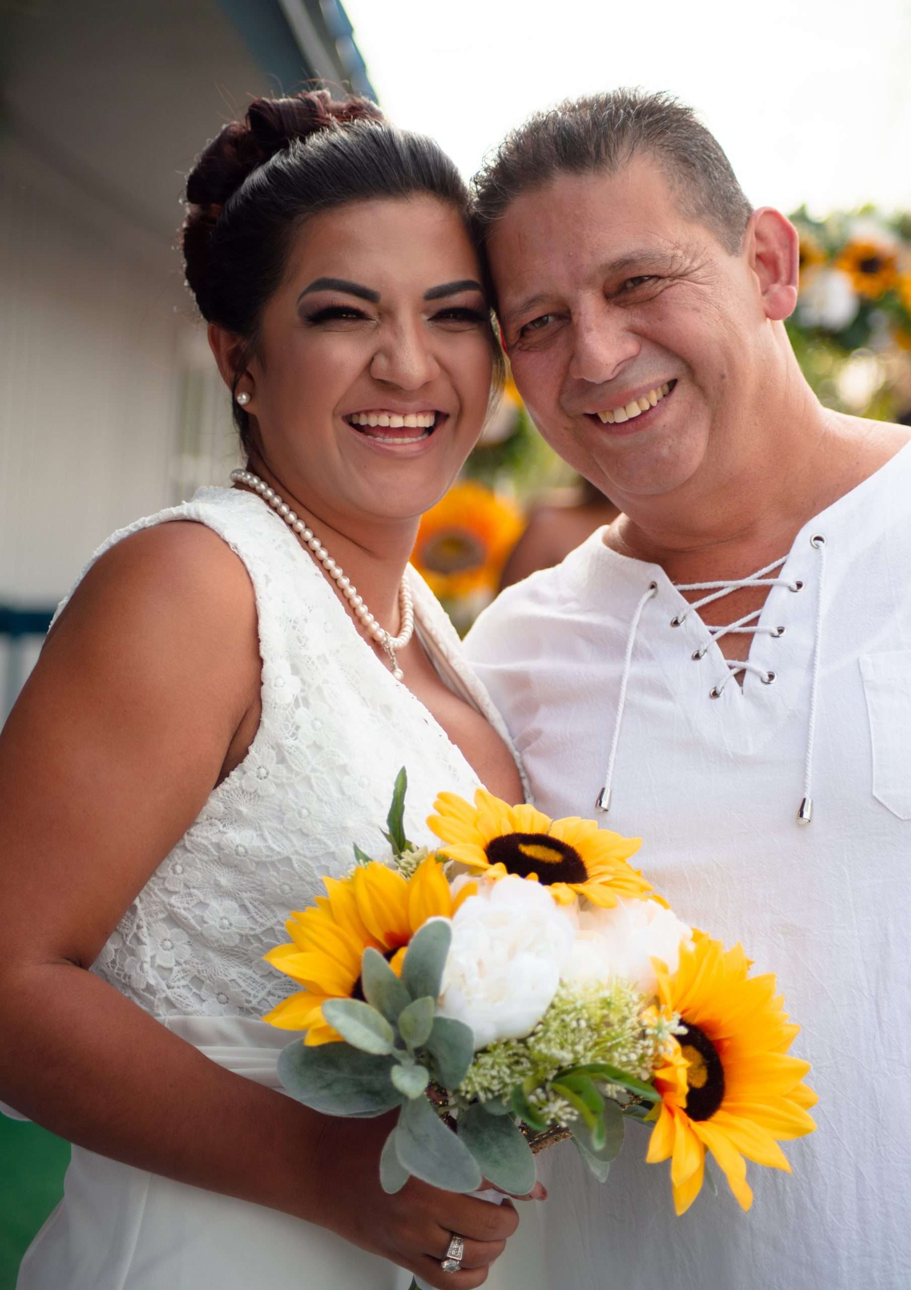 Smiling couple leaning heads together, woman in white lace dress holding a sunflower and white peony bouquet