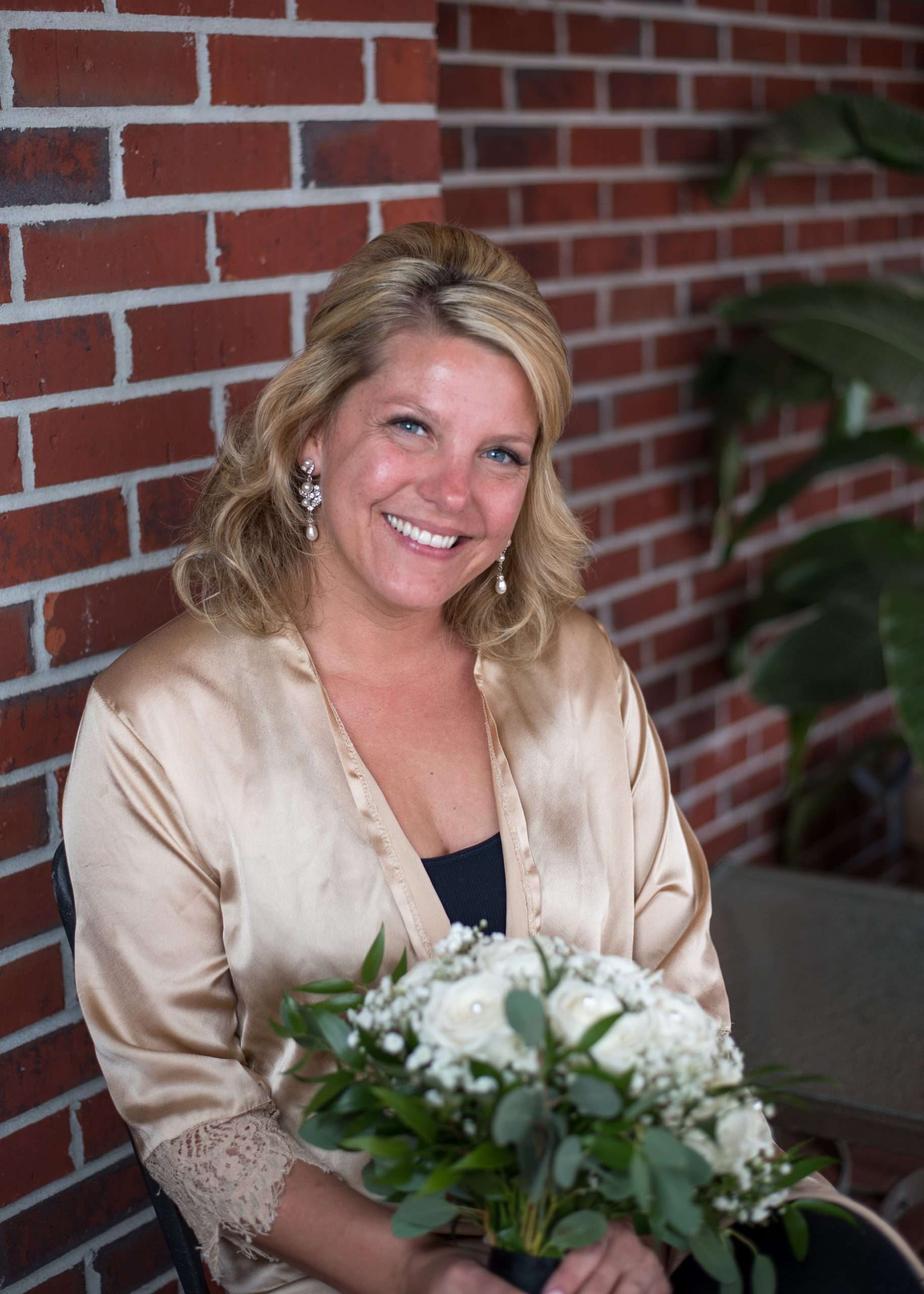 Smiling woman in a champagne satin robe holding a white rose and greenery bouquet in front of a red brick wall