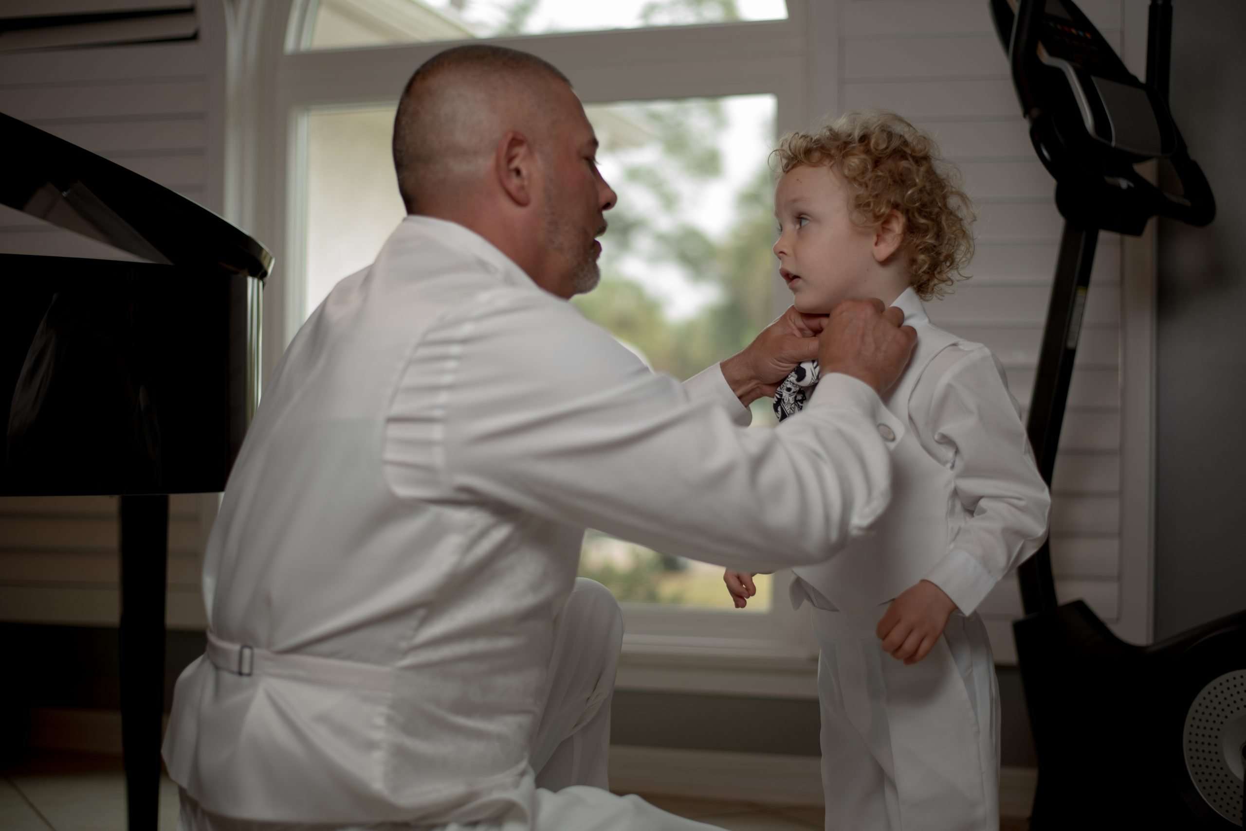 Man in a white suit adjusting a curly-haired boy's bow tie by a window