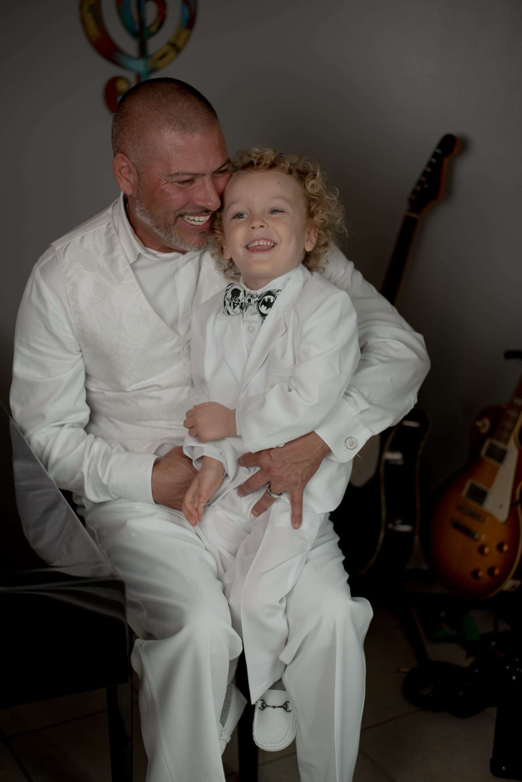Smiling man in white outfit holding a laughing curly-haired toddler in a white suit and bow tie, guitars behind them
