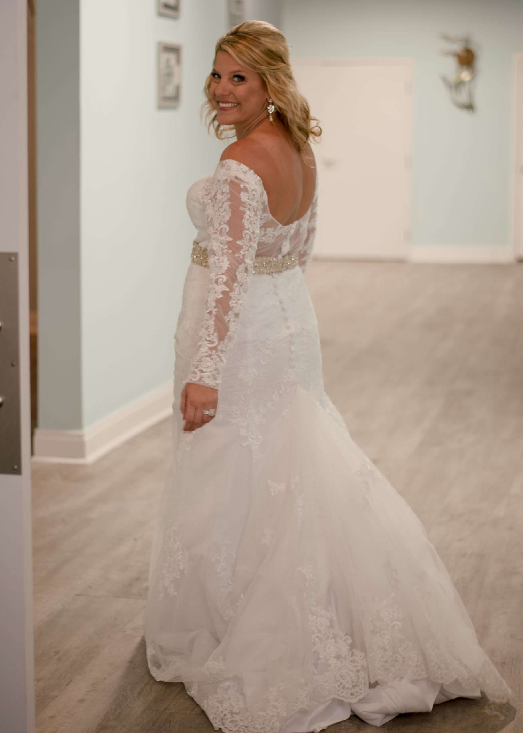 Smiling bride in a long-sleeve lace wedding gown with a beaded belt looks back over her shoulder in a hallway