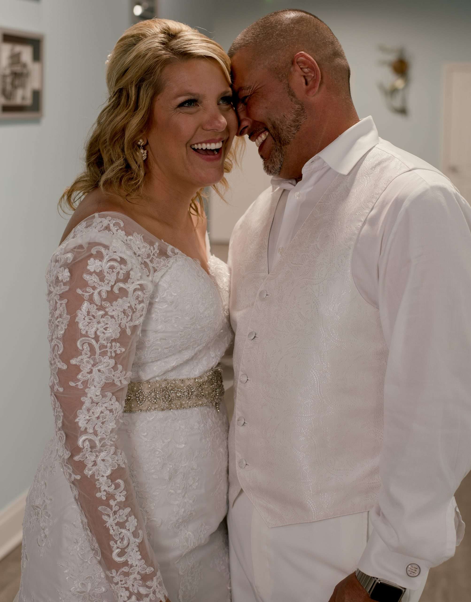 Smiling couple touching forereheads, bride in a white lace long-sleeve gown and groom in a white vest, indoors