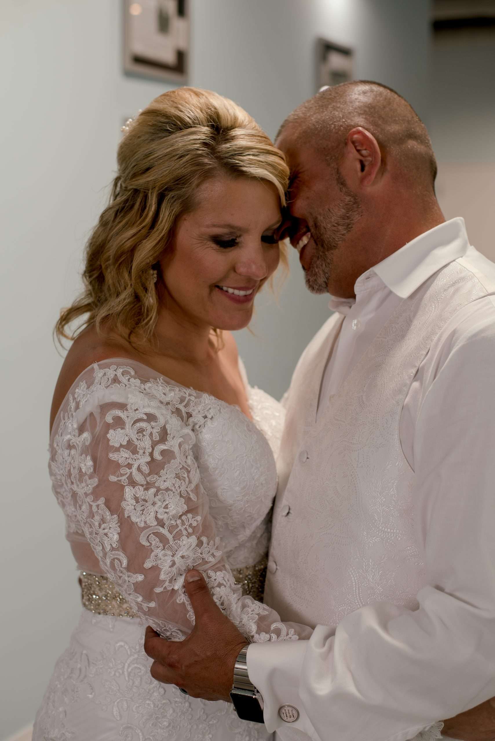 Smiling bride in a lace off-the-shoulder gown embracing her groom in a white vest and shirt, foreheads touching