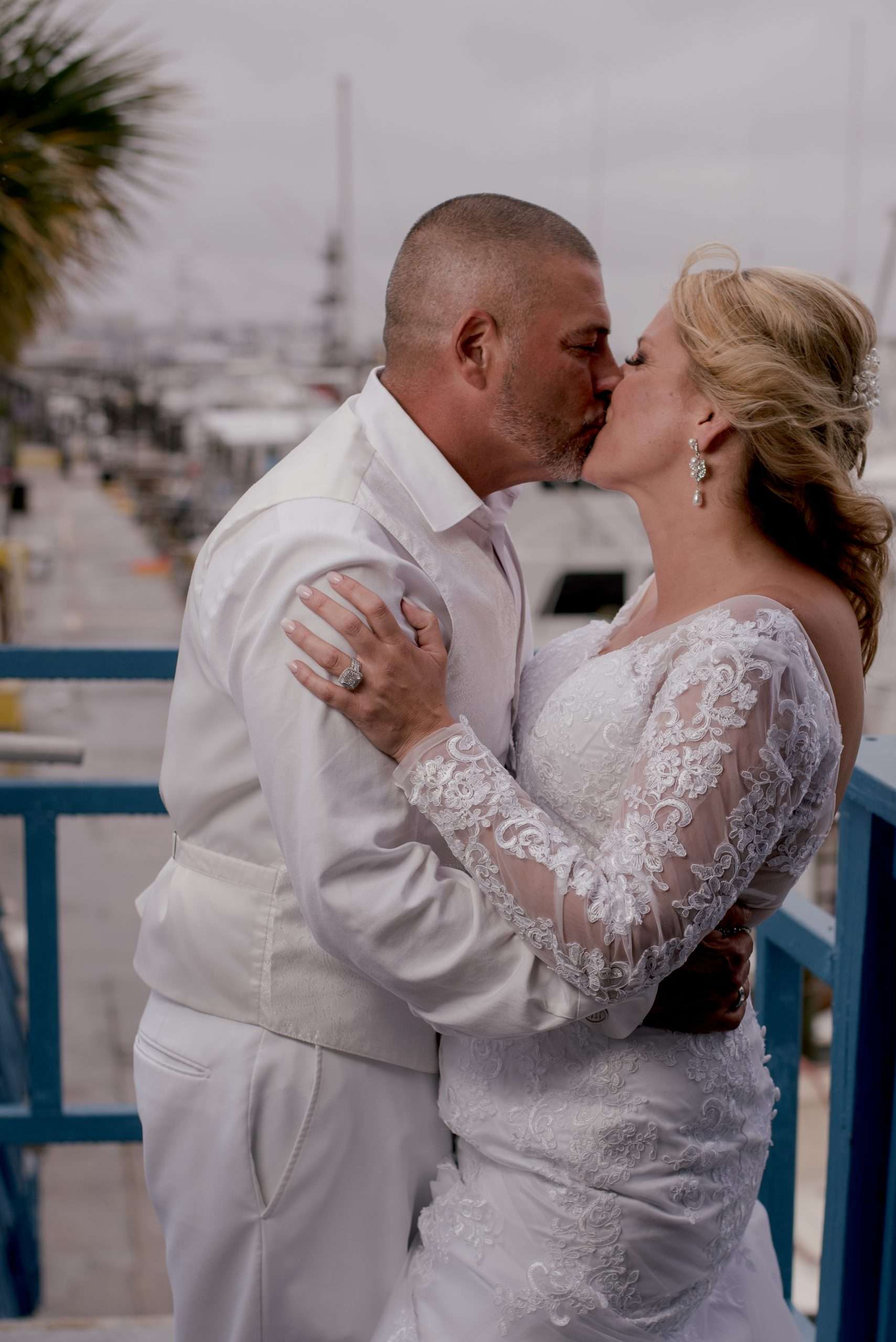 Couple in white kissing on a blue-railed balcony, bride in a long-sleeved lace gown, marina behind them
