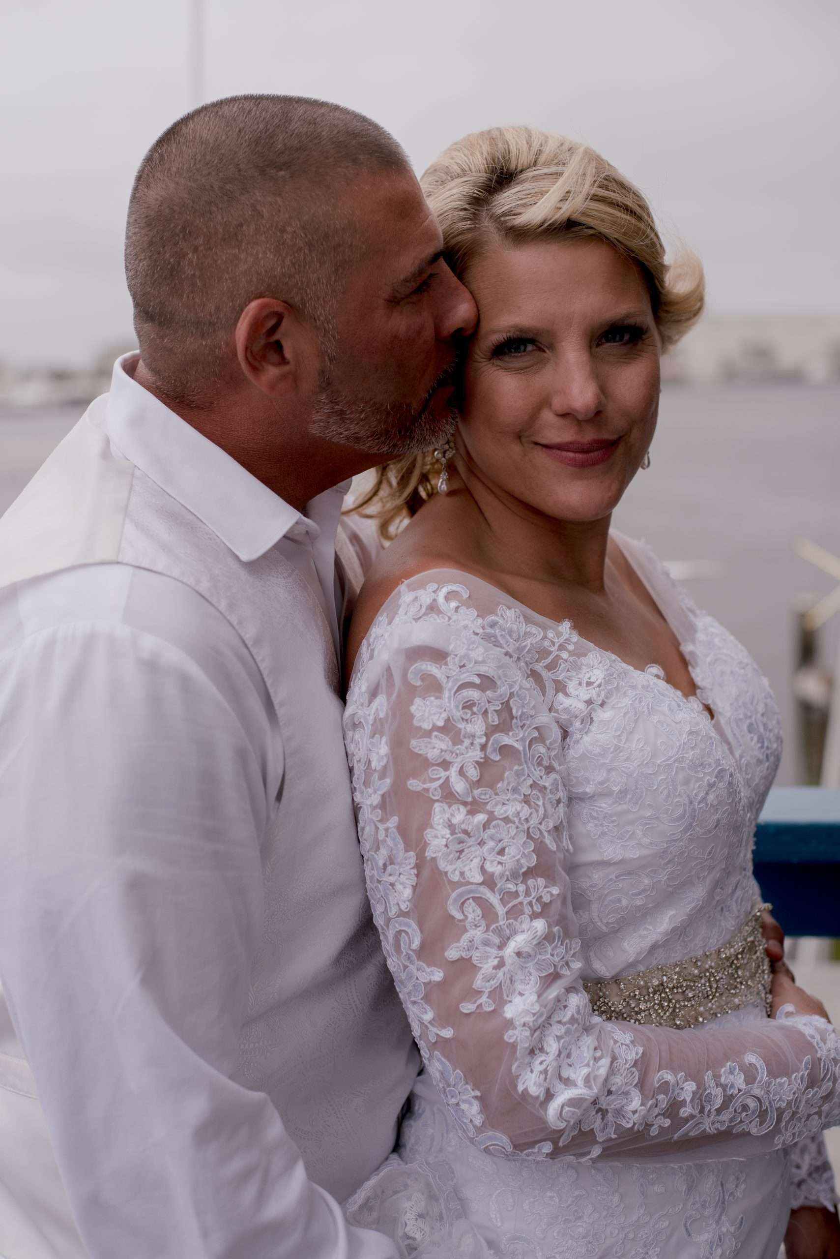 Groom in white shirt kissing his smiling bride's cheek from behind, bride in a long-sleeved white lace gown by the water