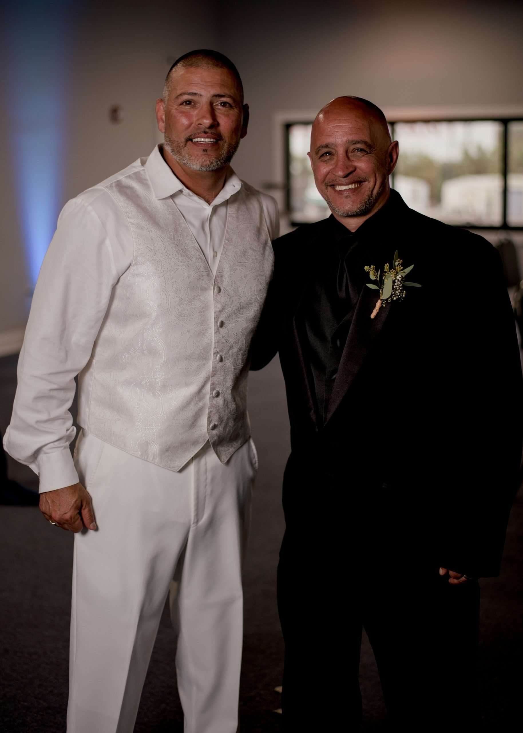 Two smiling men posing side by side at a wedding, one in a white vest and pants, the other in a black suit with boutonniere