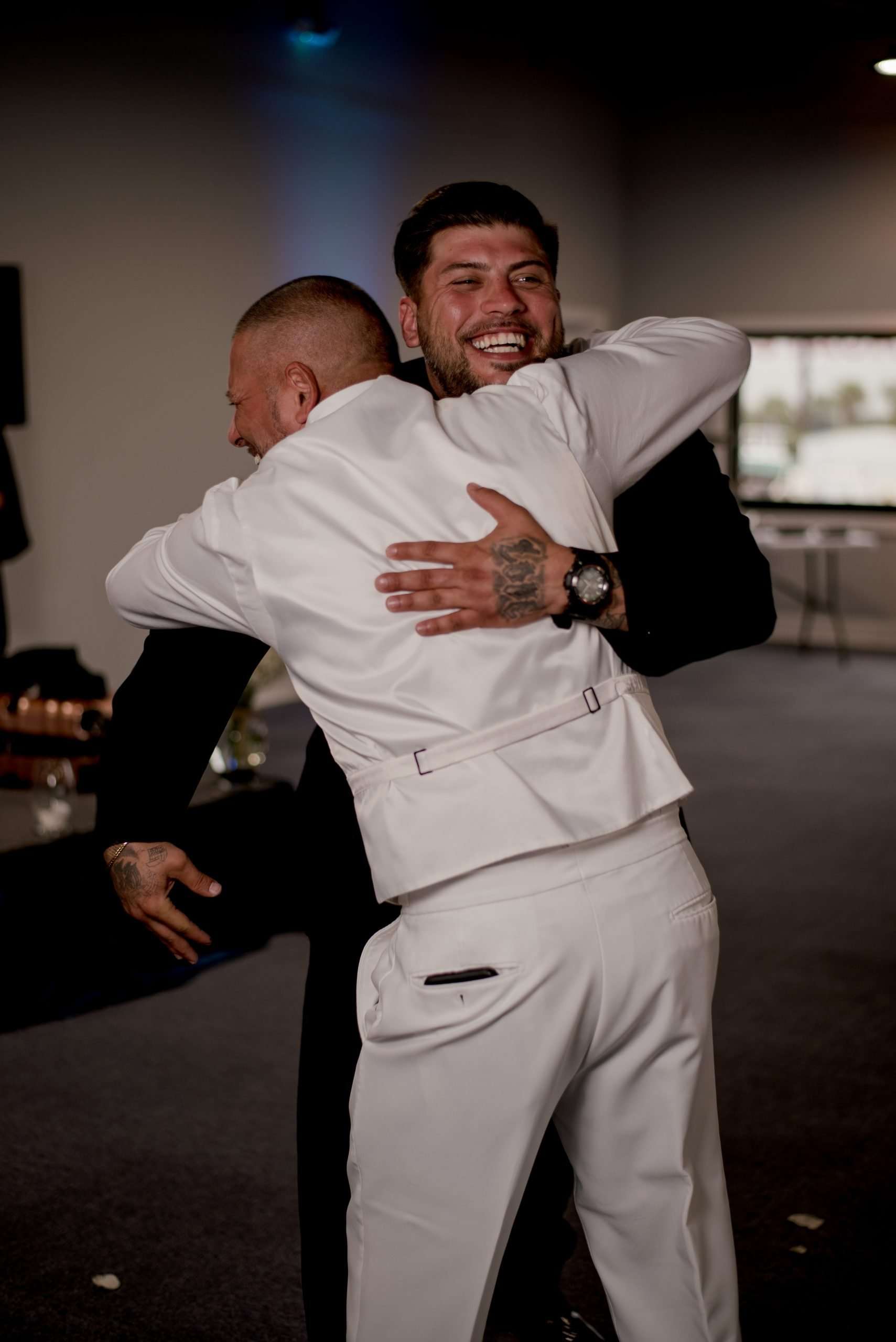 Two men in a warm embrace, one in a white vest and trousers, the other in black laughing with his eyes closed