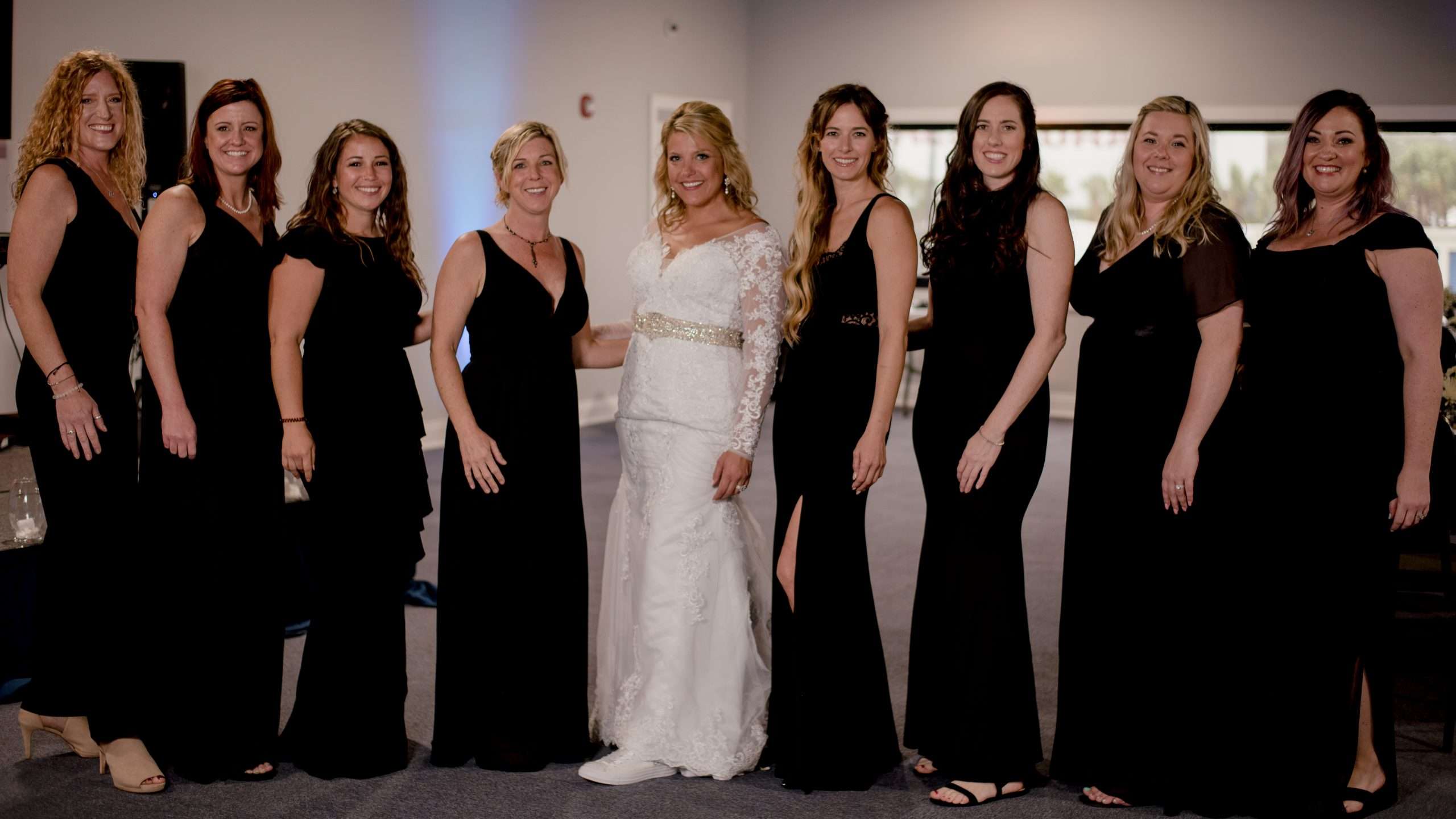 Bride in a white lace gown smiling, surrounded by eight bridesmaids in long black dresses indoors