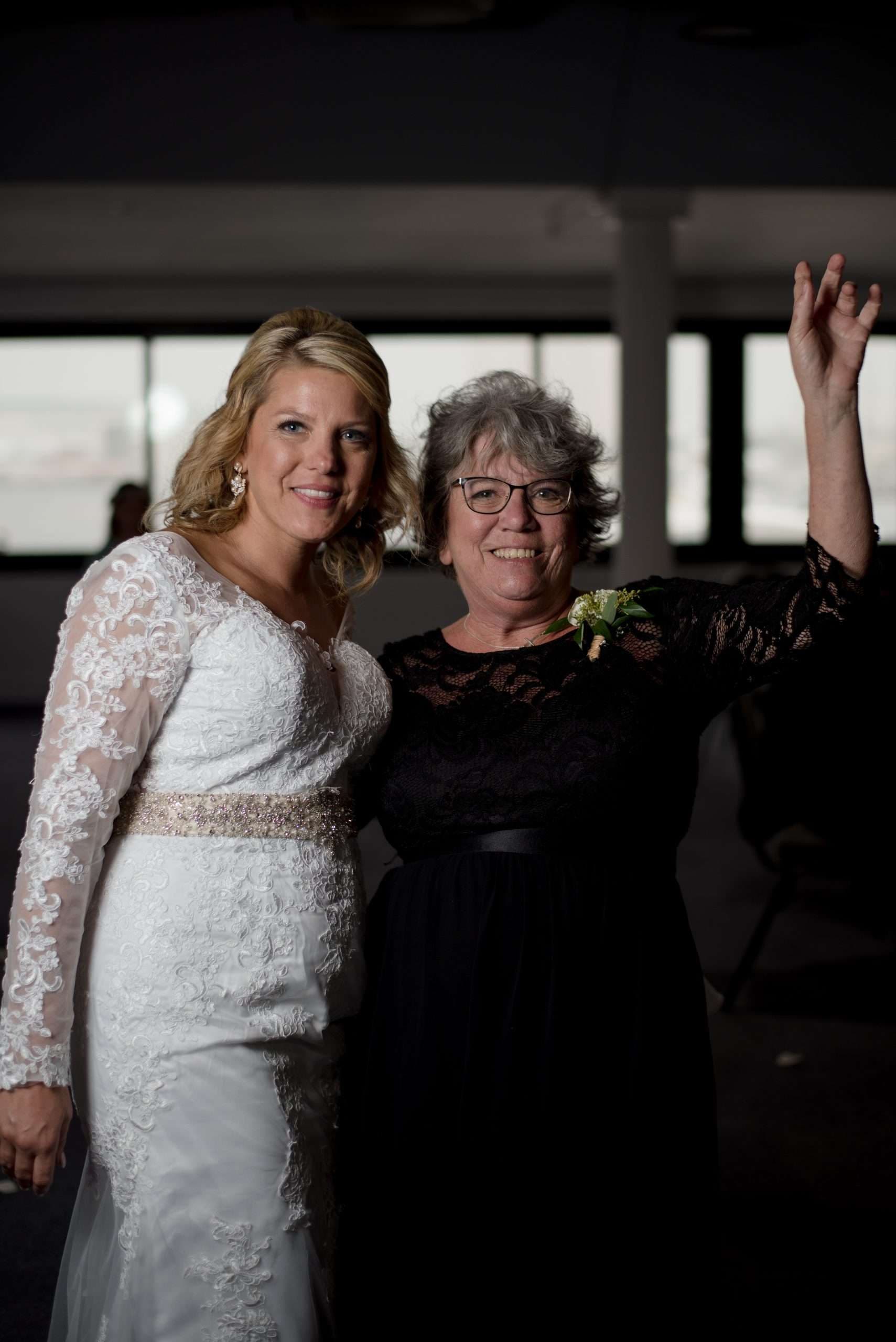 Bride in a white lace gown smiling beside an older woman in a black lace dress who raises one hand