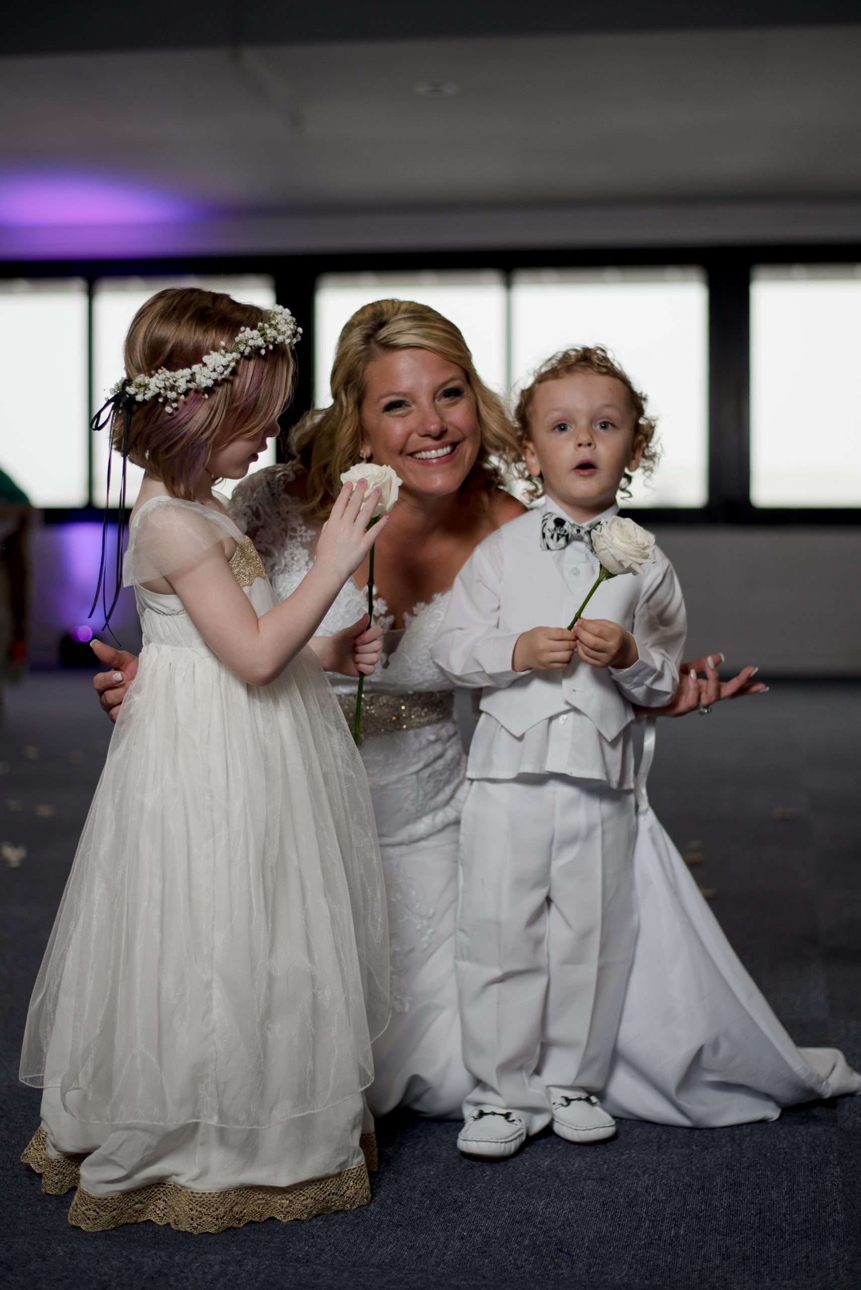 Smiling bride in a white lace gown with a flower girl in a floral crown and a ring bearer holding a white rose