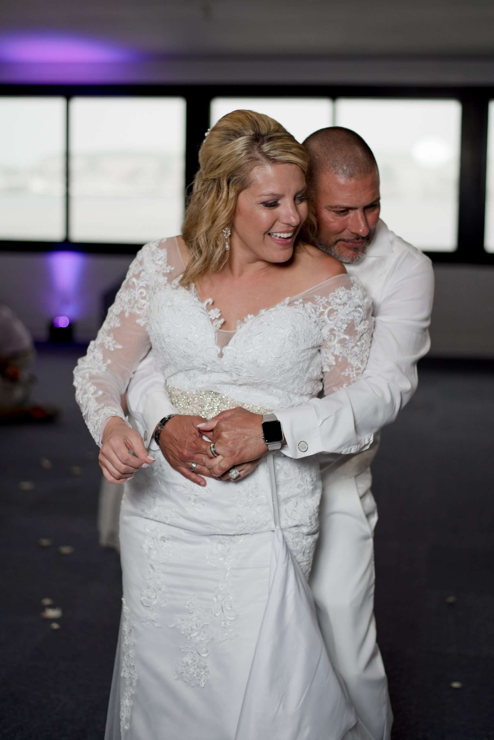 Groom in white embracing his laughing bride from behind during a dance, she wears a long-sleeve lace wedding gown