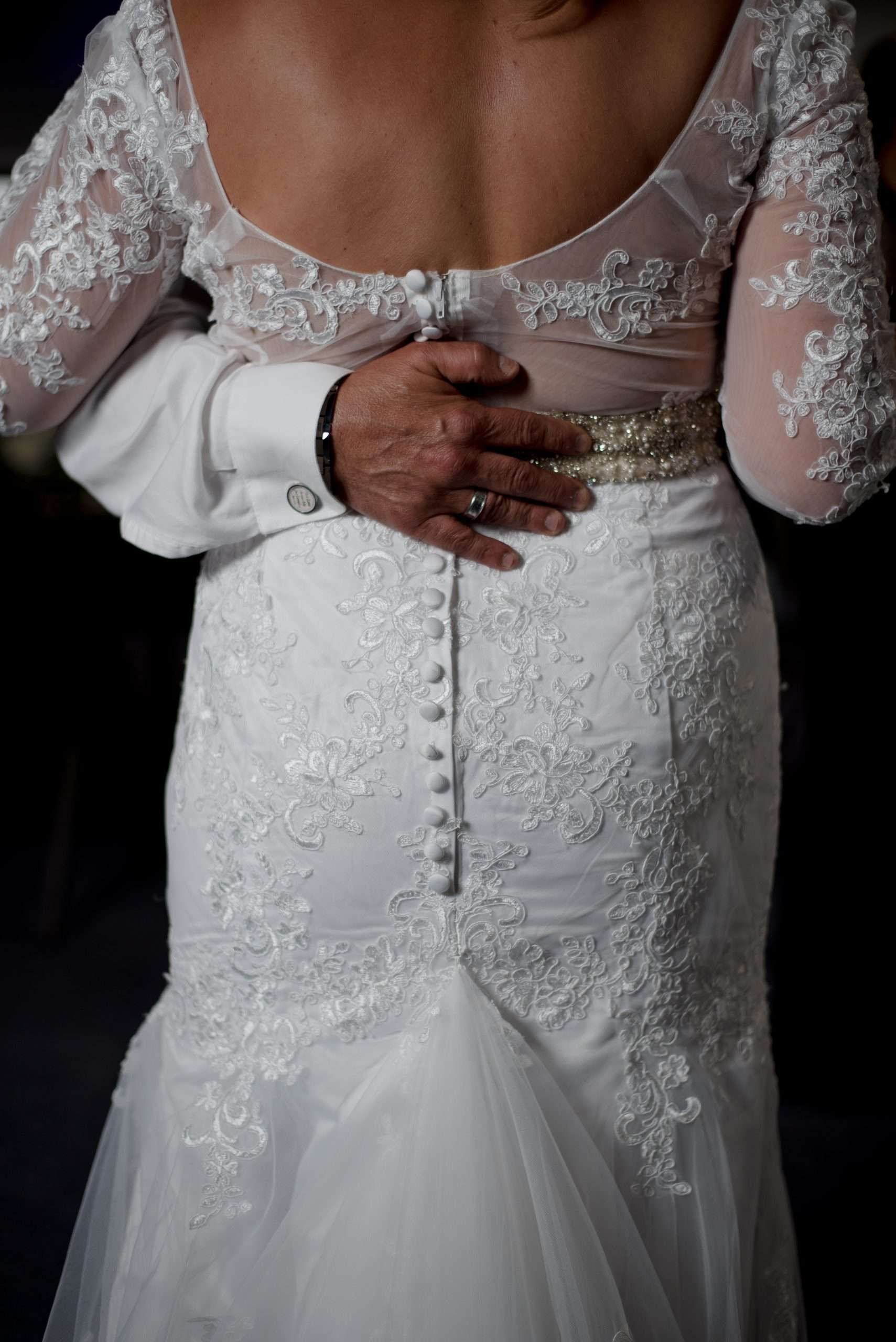 Close-up of a hand resting on the back of a bride in a lace gown with buttons and a beaded belt
