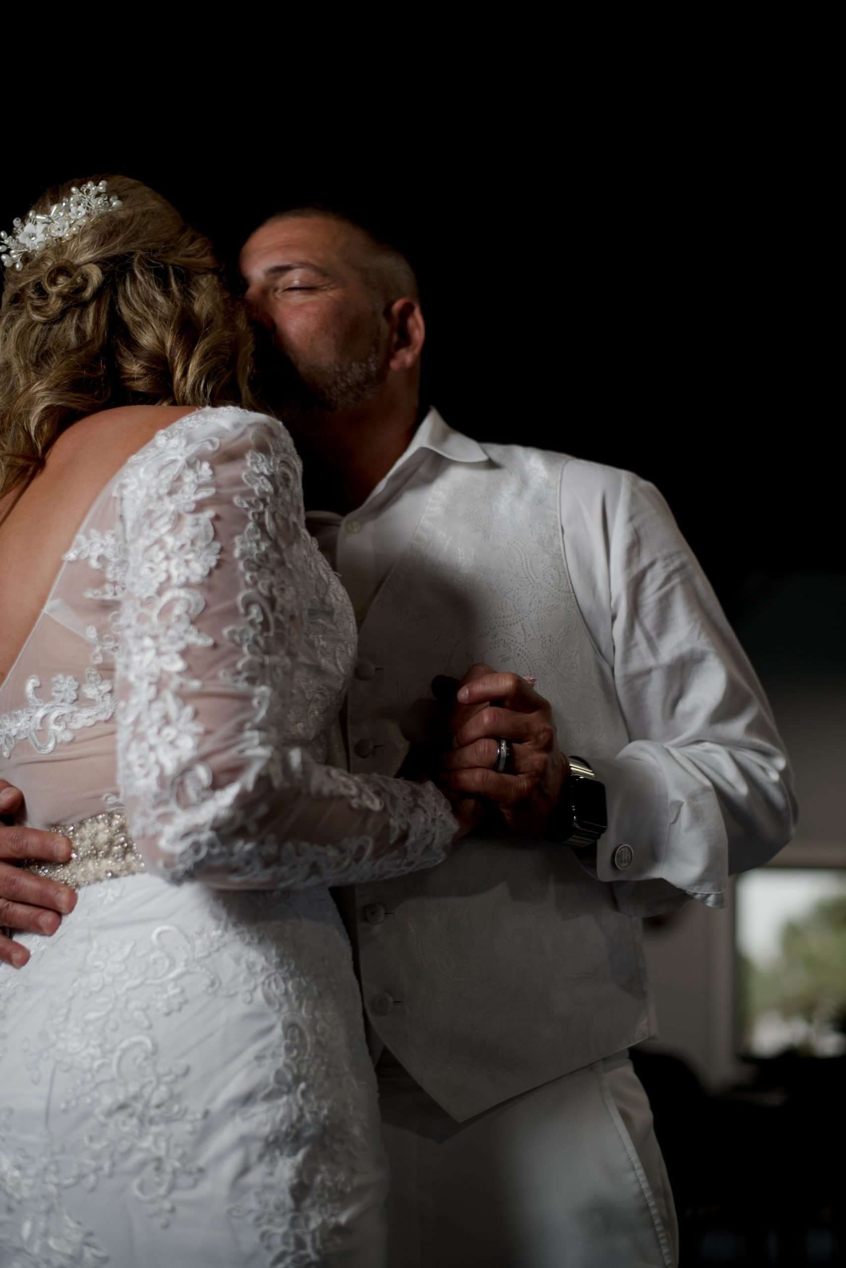 Groom in white vest kissing his bride's temple during a slow dance, she wears a lace gown and beaded hair piece