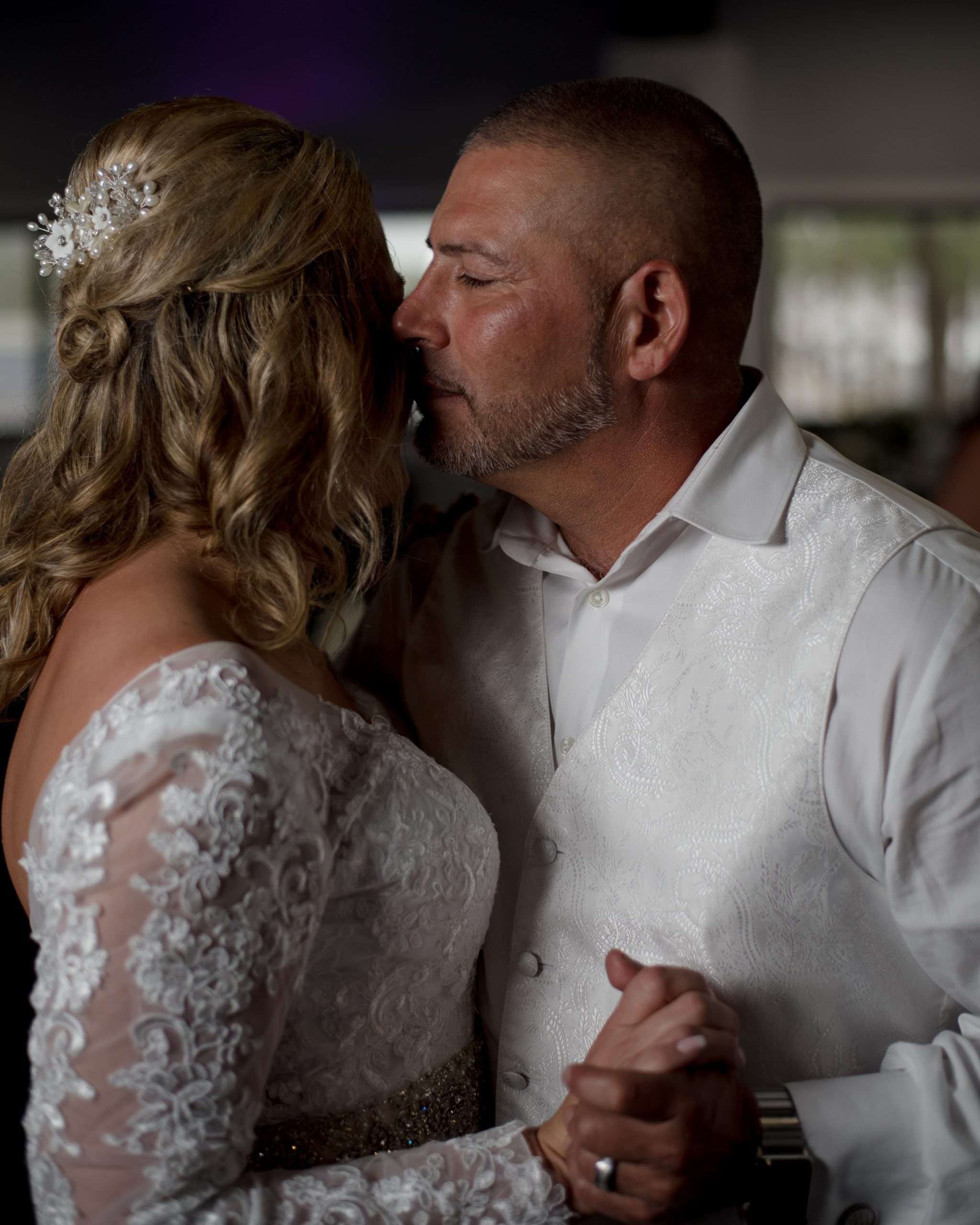 Groom in white vest kissing his bride's forehead during a slow dance, her lace sleeve and hair comb visible