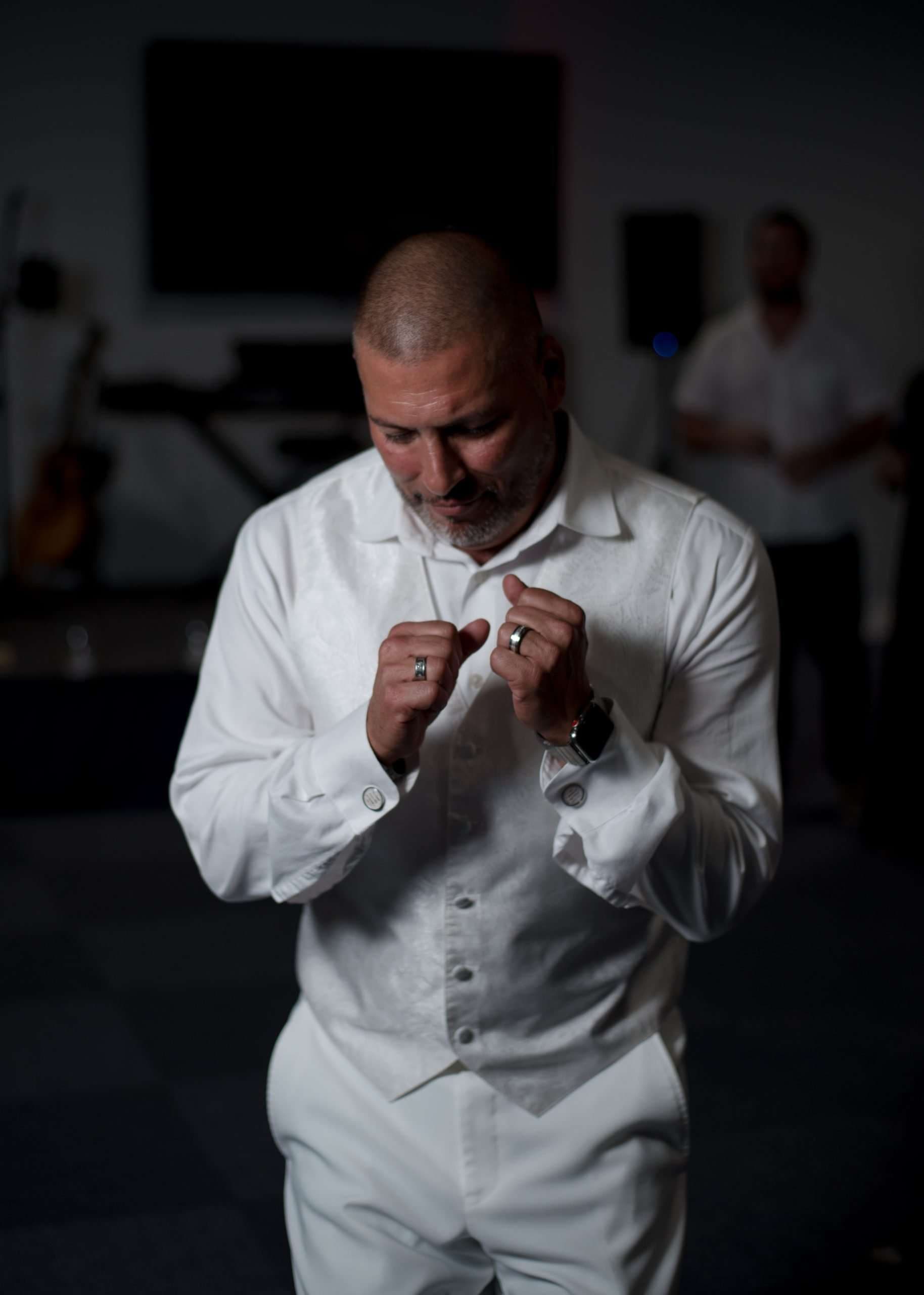 Man in a white vest and shirt looking down with fists raised near his chest in a dimly lit room