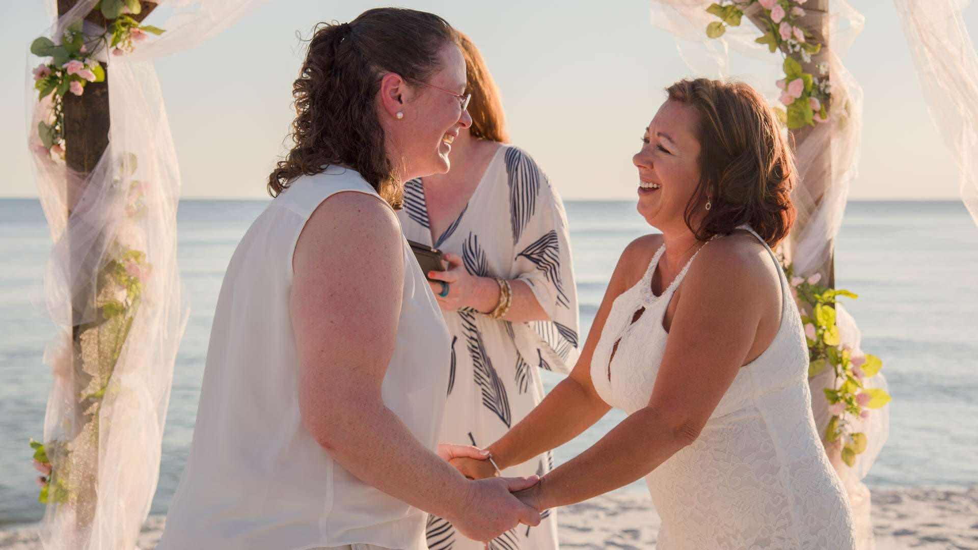 Two women in white laughing and holding hands under a flower-draped tulle arch on the beach, officiant behind them