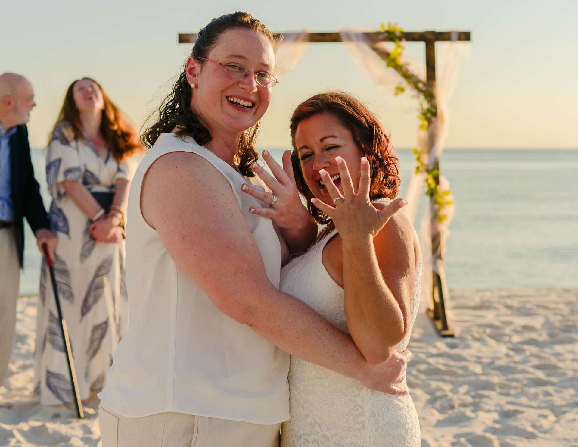 Two smiling women embrace on the beach at sunset, one showing her ring, with a floral wedding arch behind them