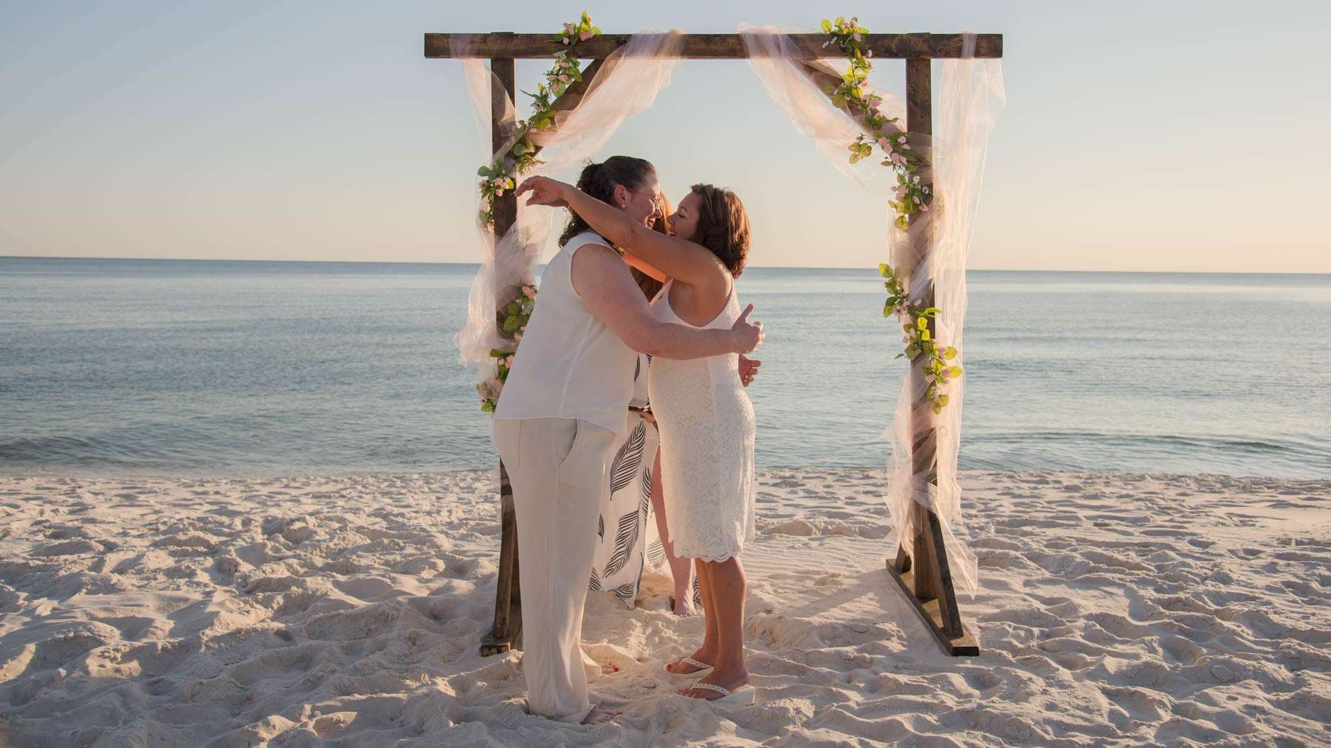 Couple embracing under a wooden beach wedding arch draped with sheer fabric and greenery at sunset