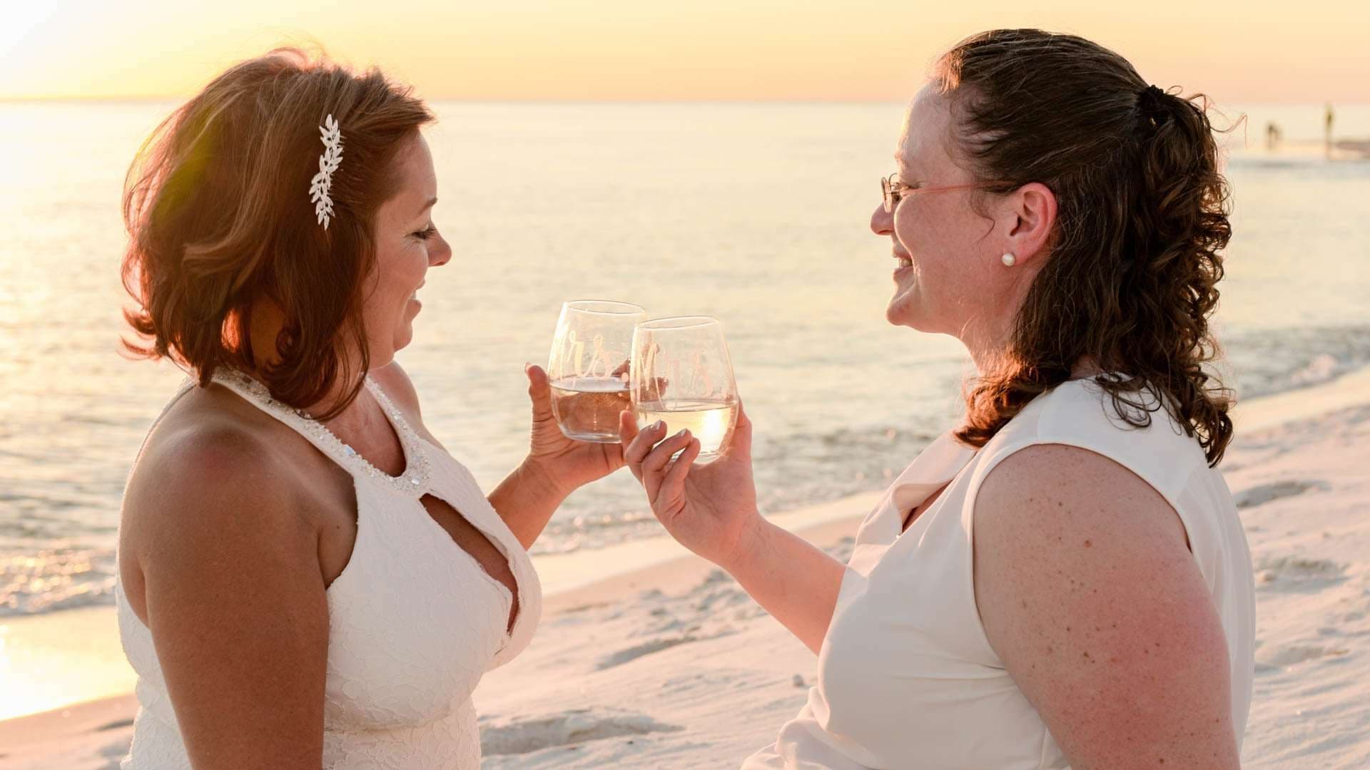Two women in white laughing and toasting with wine glasses on the beach at sunset
