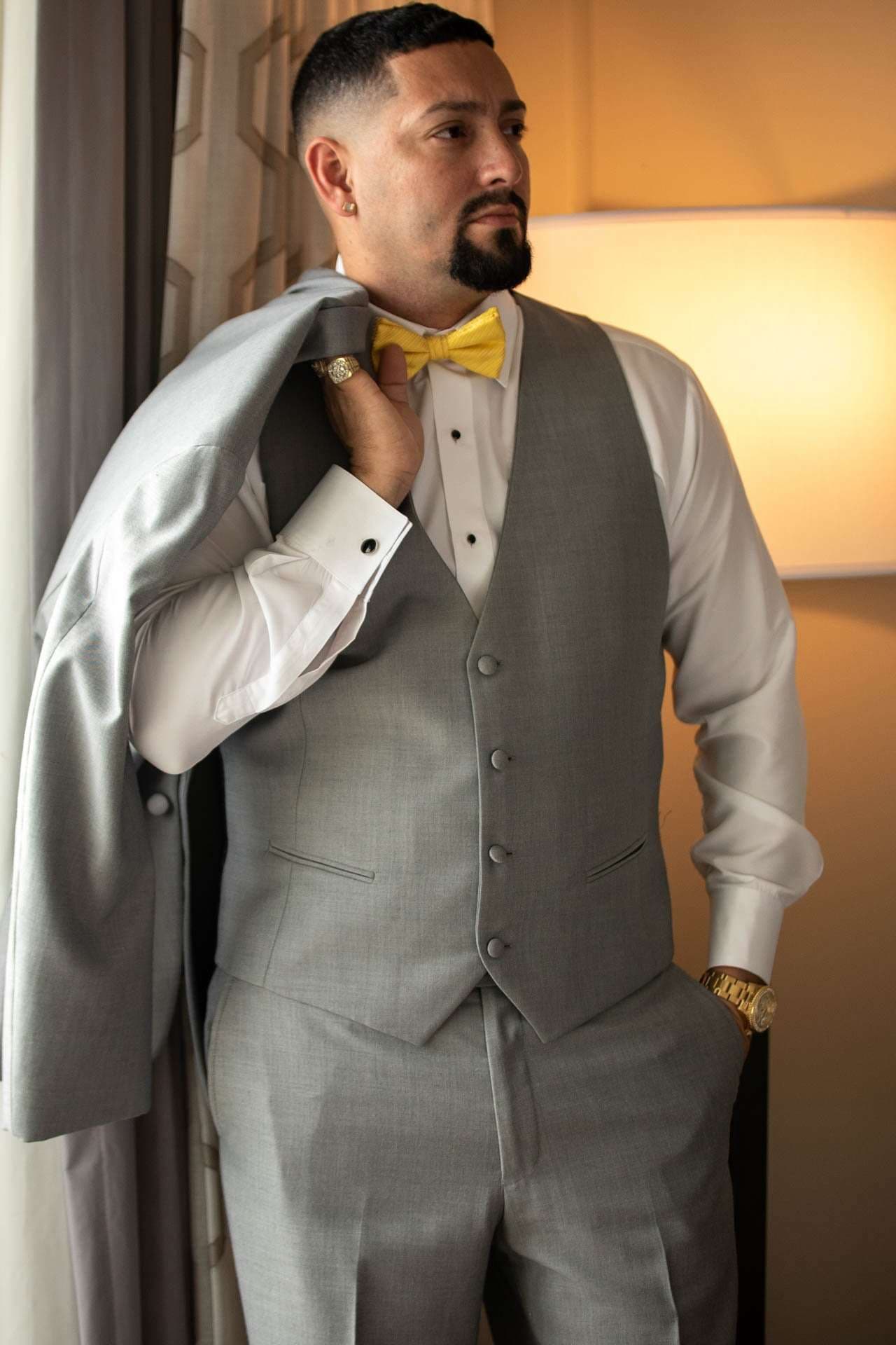 Groom in gray vest and yellow bow tie with suit jacket slung over his shoulder, gazing aside beside a lamp