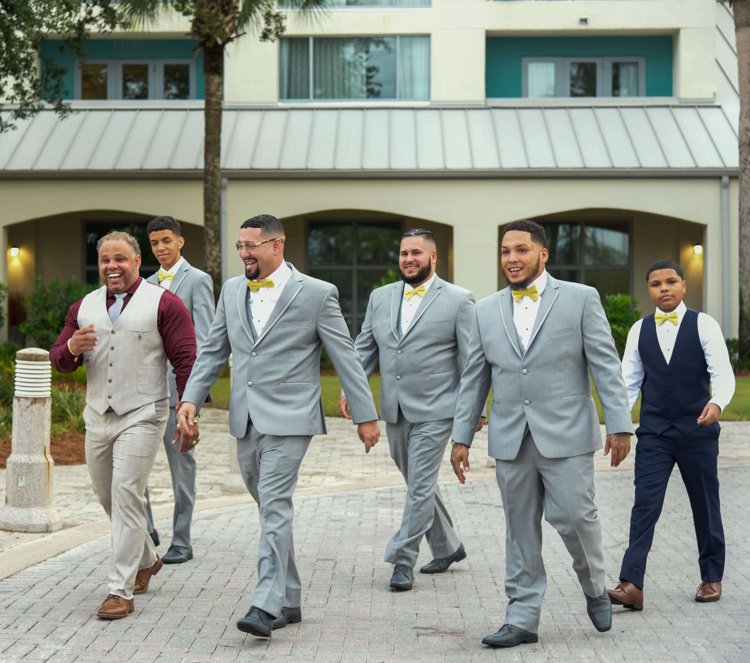 Five men in gray suits and yellow bow ties walking and laughing together along a brick paved path outside a hotel