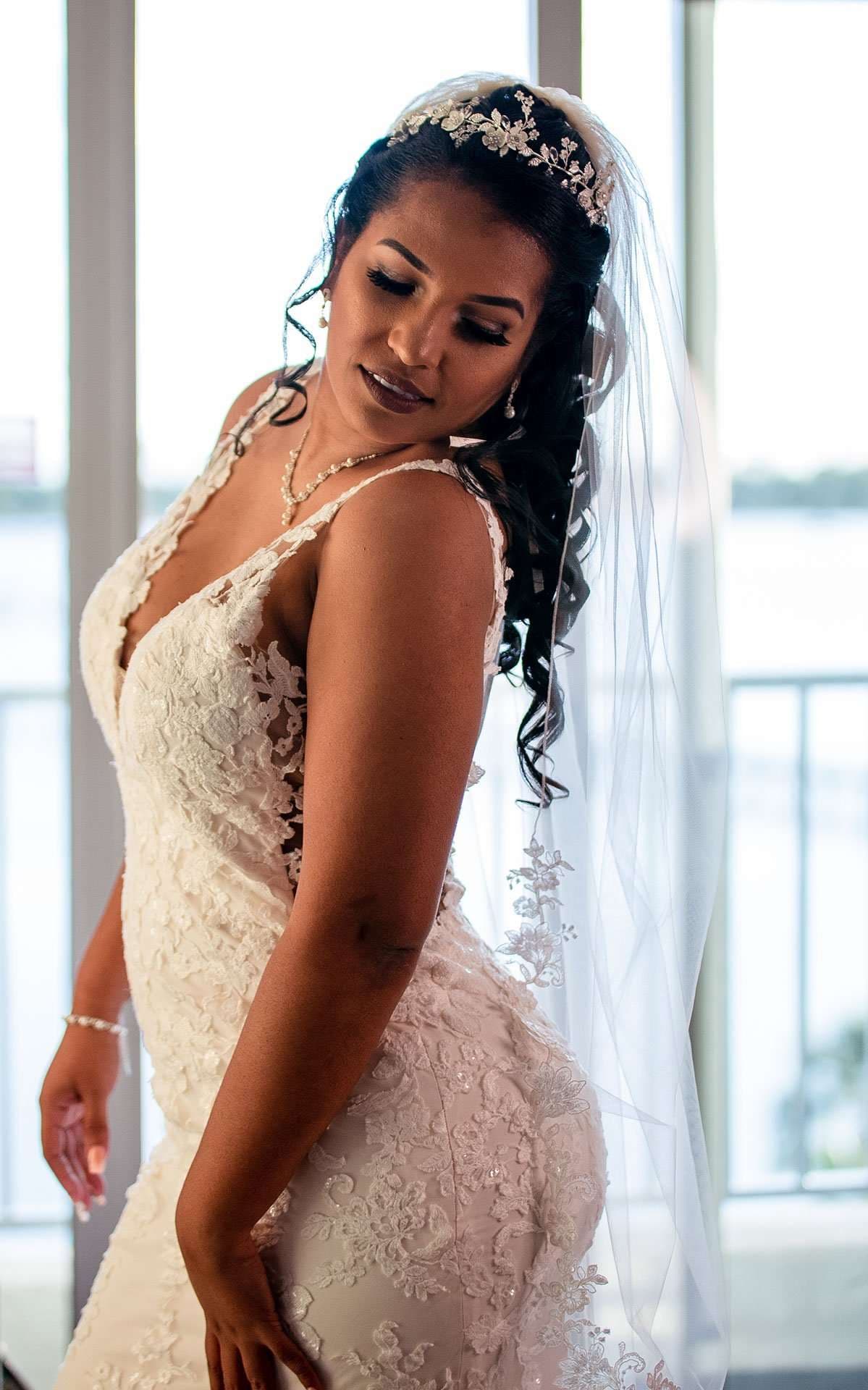Bride in a white lace gown and long veil glances over her shoulder beside bright windows