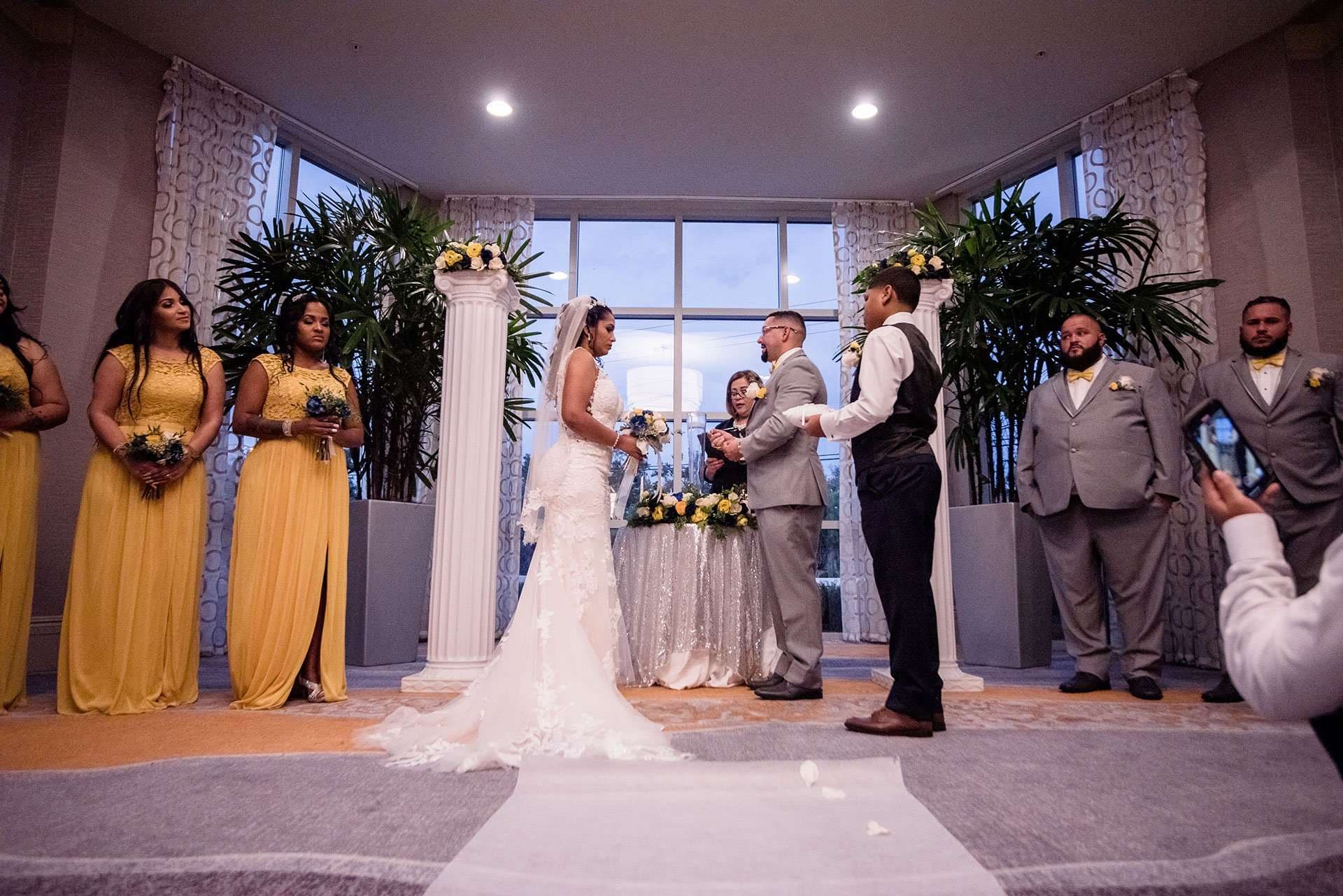Couple exchanging vows during an indoor ceremony, with bridesmaids in yellow gowns and groomsmen in gray suits looking on