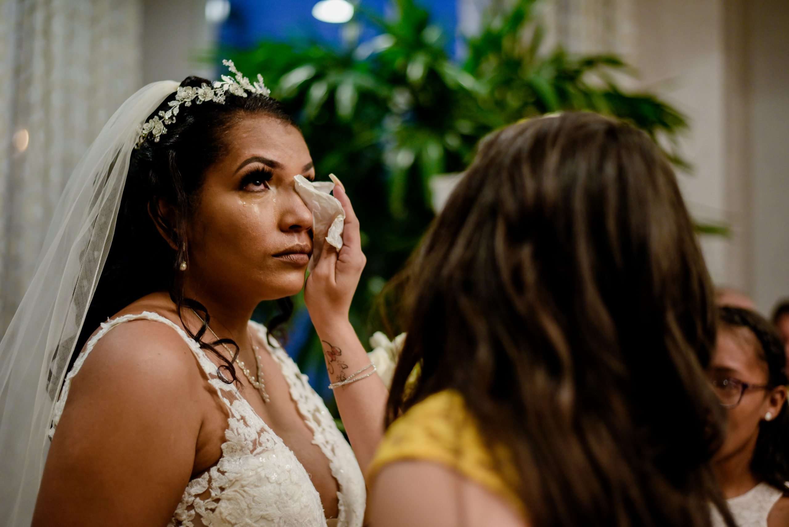 Tearful bride in a lace gown and floral headpiece dabs her eye with a tissue while a guest faces her