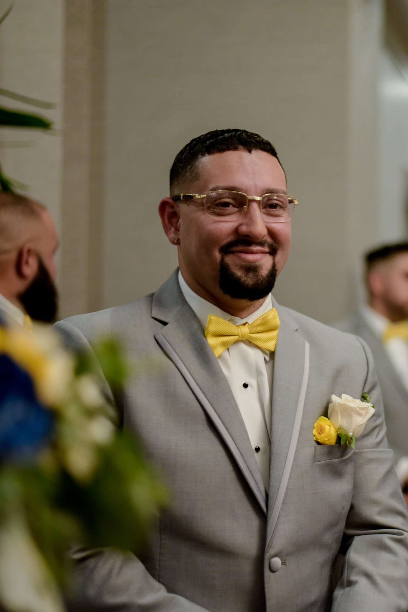 Smiling man in a gray suit with yellow bow tie and rose boutonniere, glasses, and goatee at a wedding