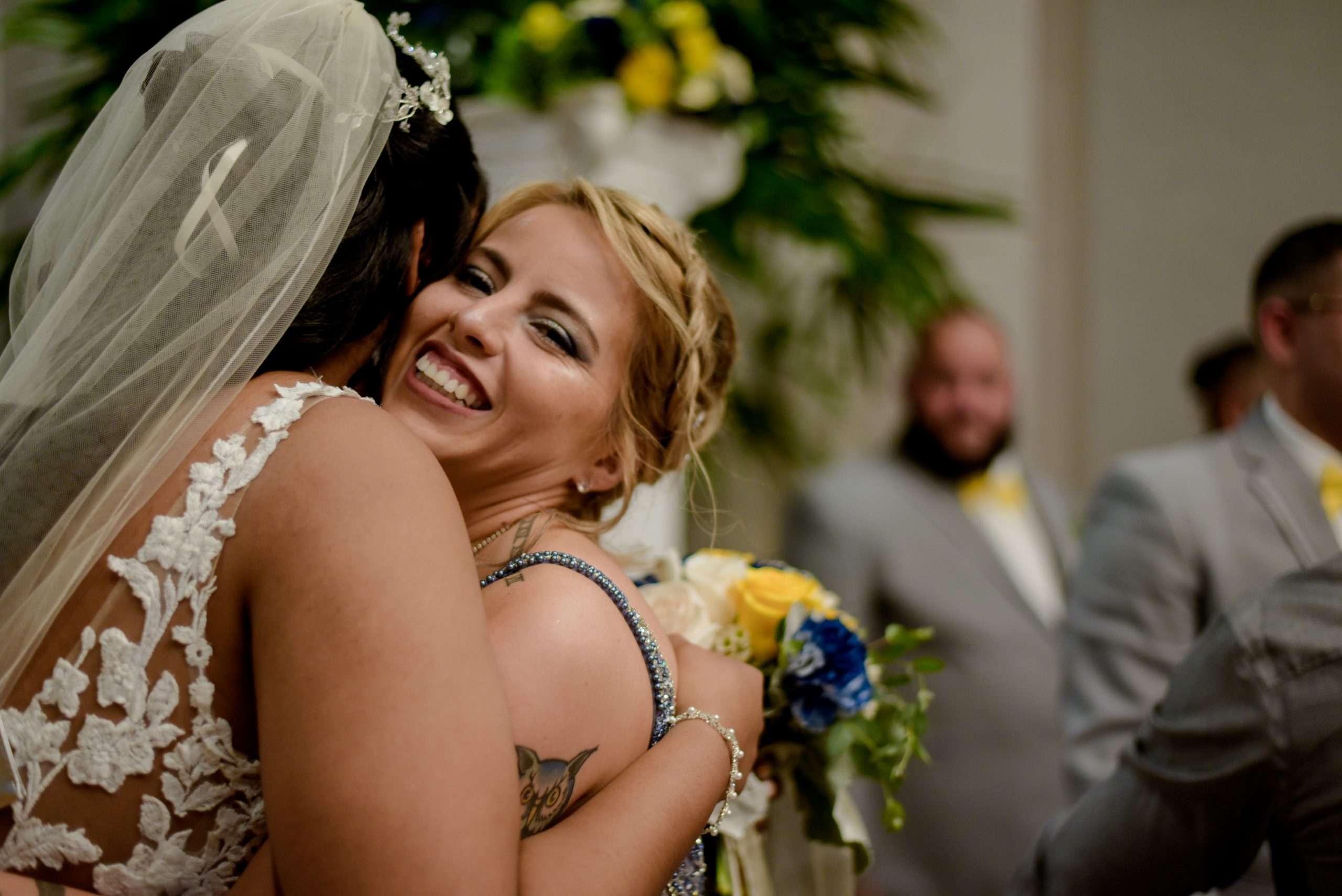 Bride in lace gown and veil hugging a smiling blonde guest beside a yellow and blue bouquet