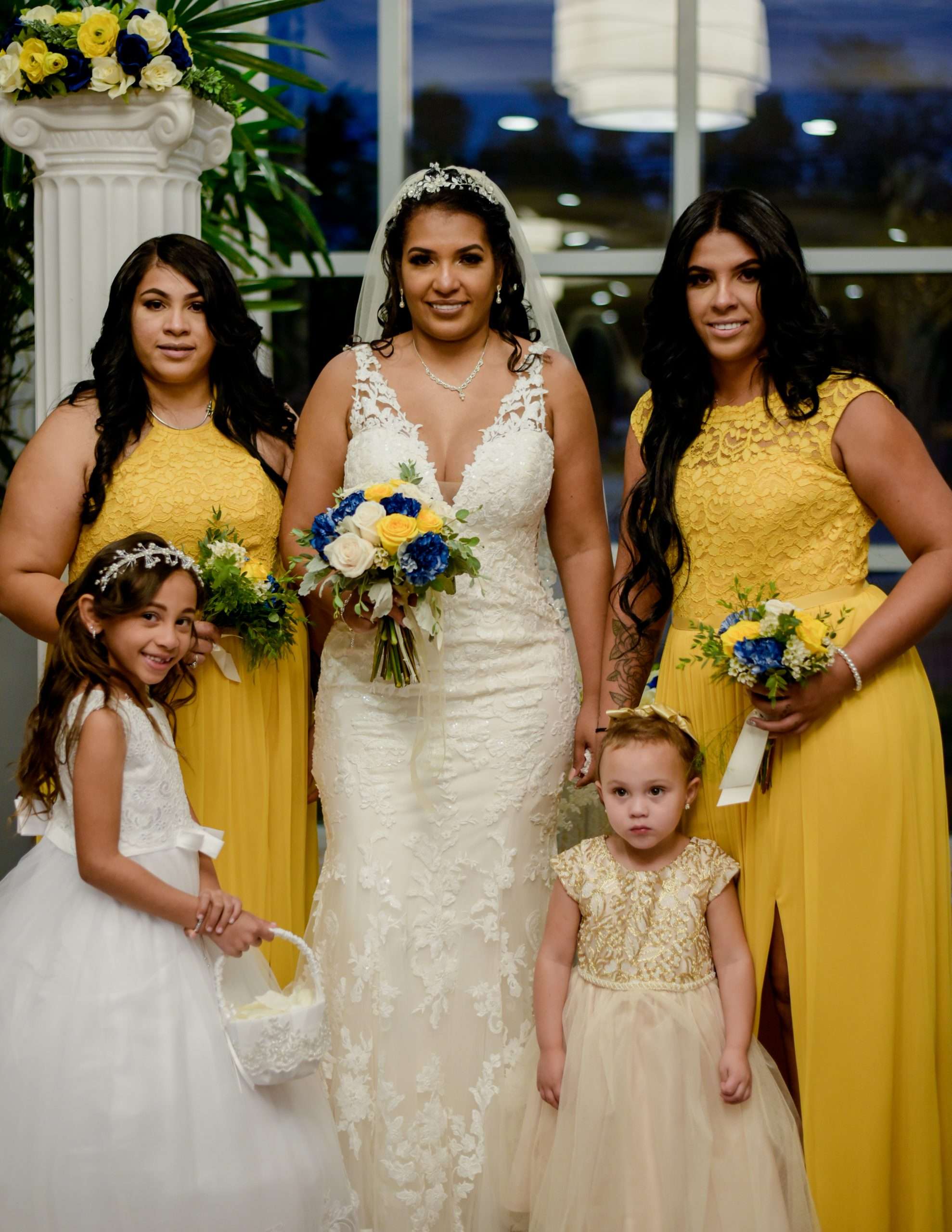 Bride in a white lace gown with two bridesmaids in yellow and two flower girls, holding blue and yellow bouquets