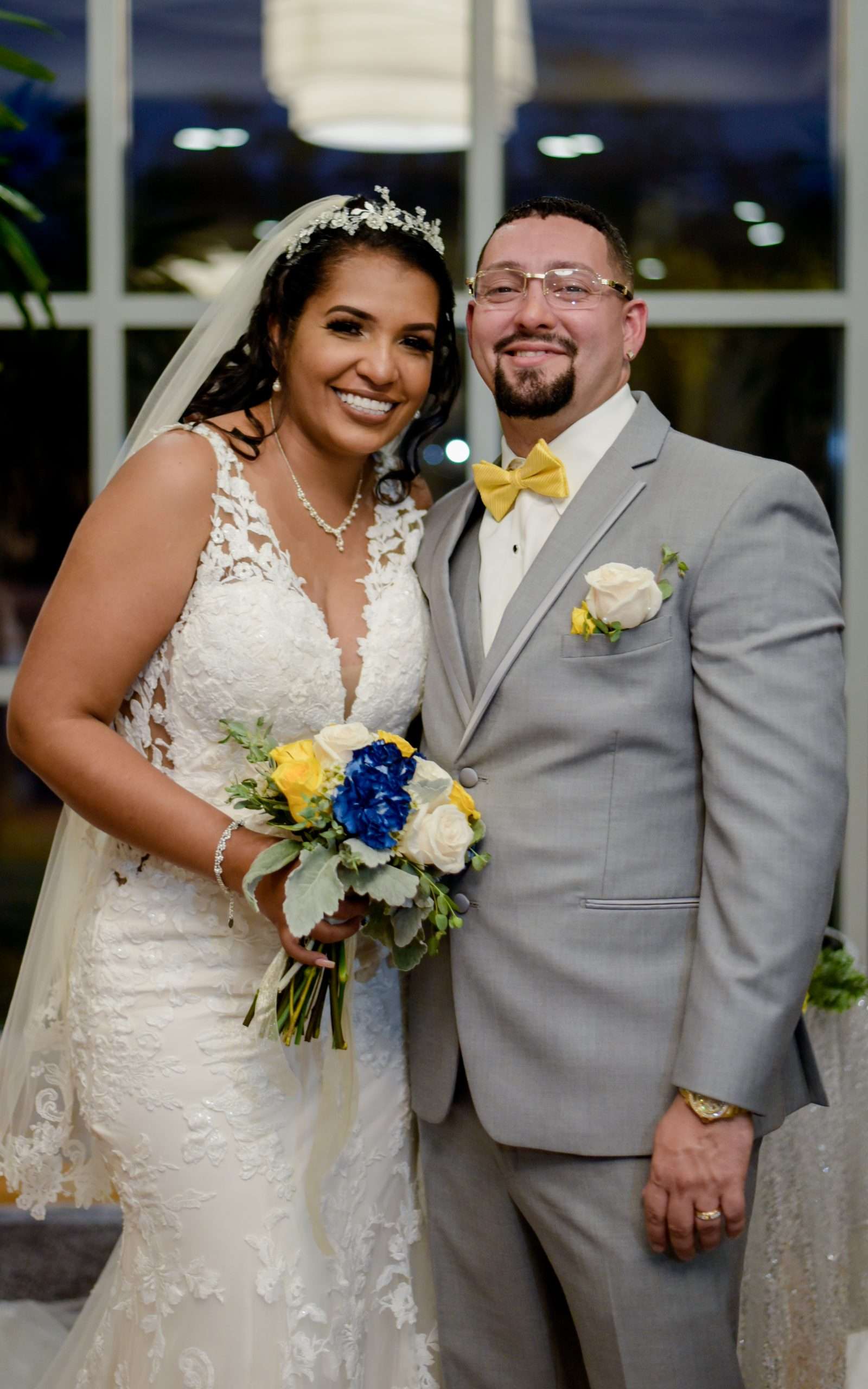 Smiling couple posing together, bride in a lace gown holding a blue and yellow bouquet, groom in gray suit and yellow bow tie
