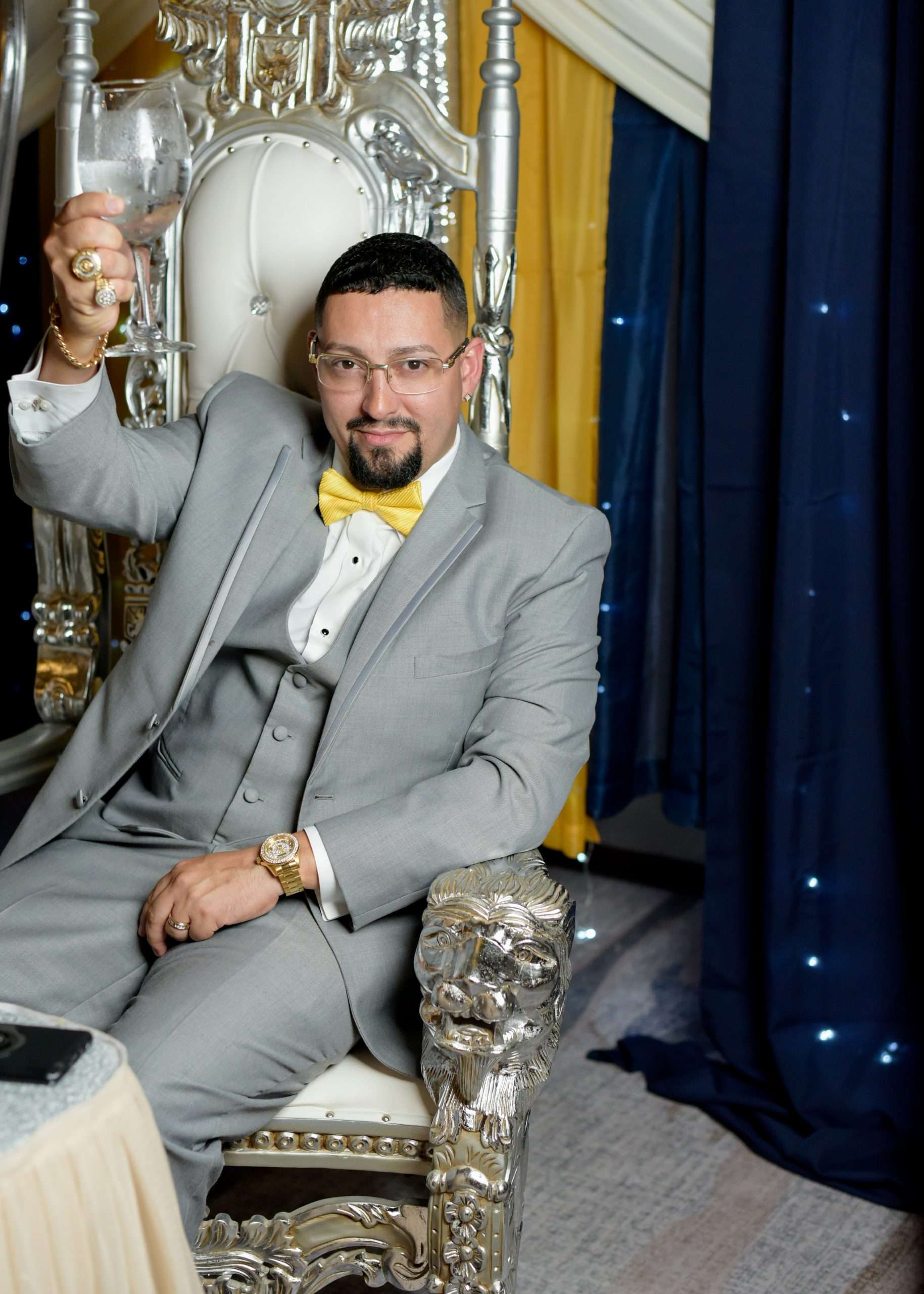 Man in gray three-piece suit and yellow bow tie raising a glass while seated on an ornate silver throne chair