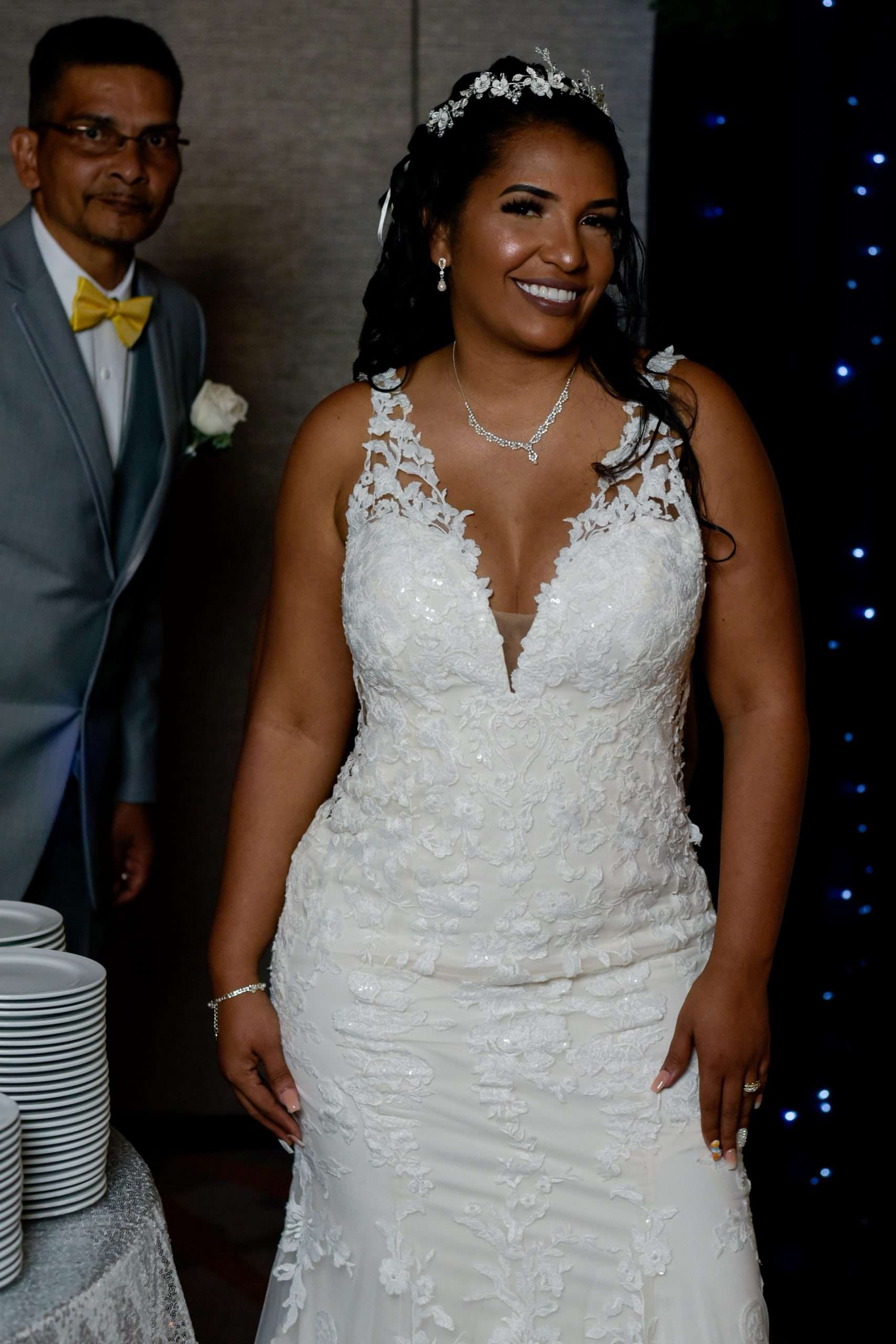 Smiling bride in a white lace gown and tiara, with a man in a gray suit behind her