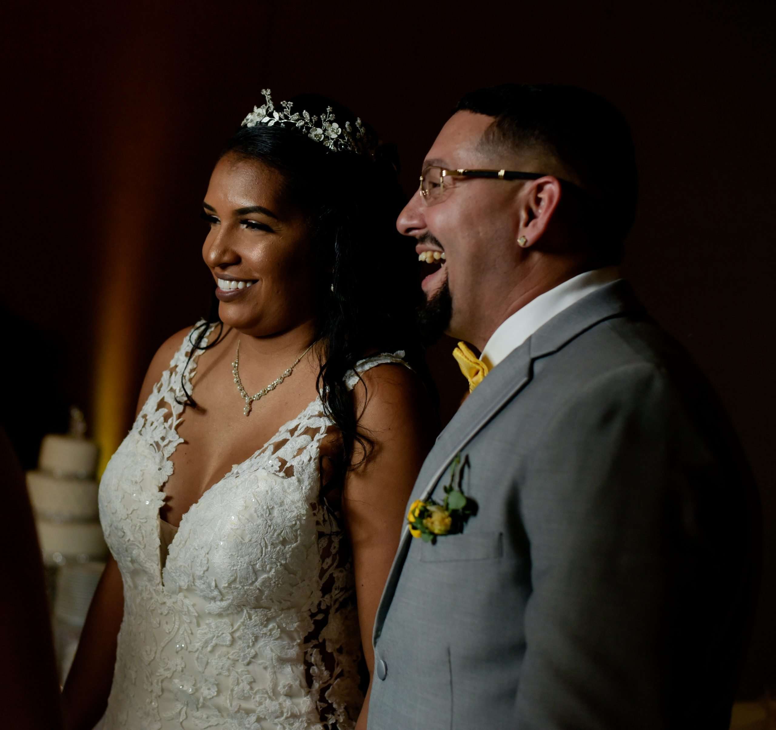 Smiling bride in a lace gown and tiara beside a laughing groom in a gray suit with yellow bow tie