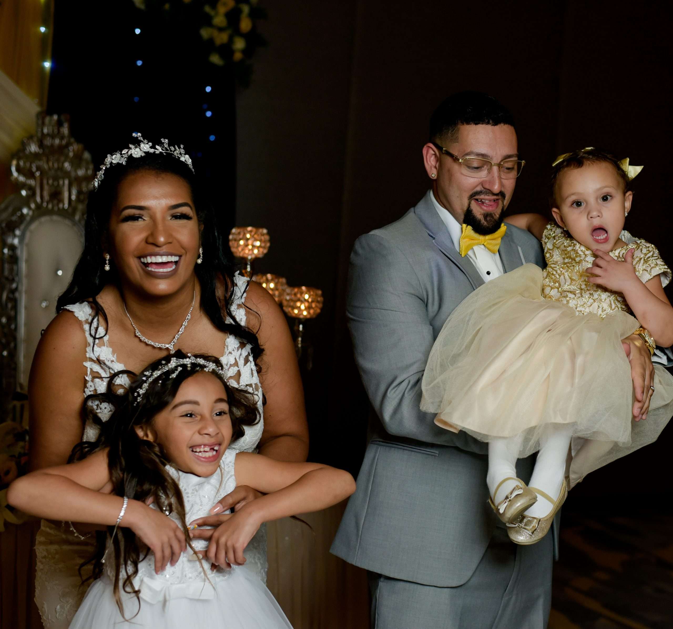Laughing bride in a lace gown and tiara with two flower girls, while the groom in a gray suit holds a toddler