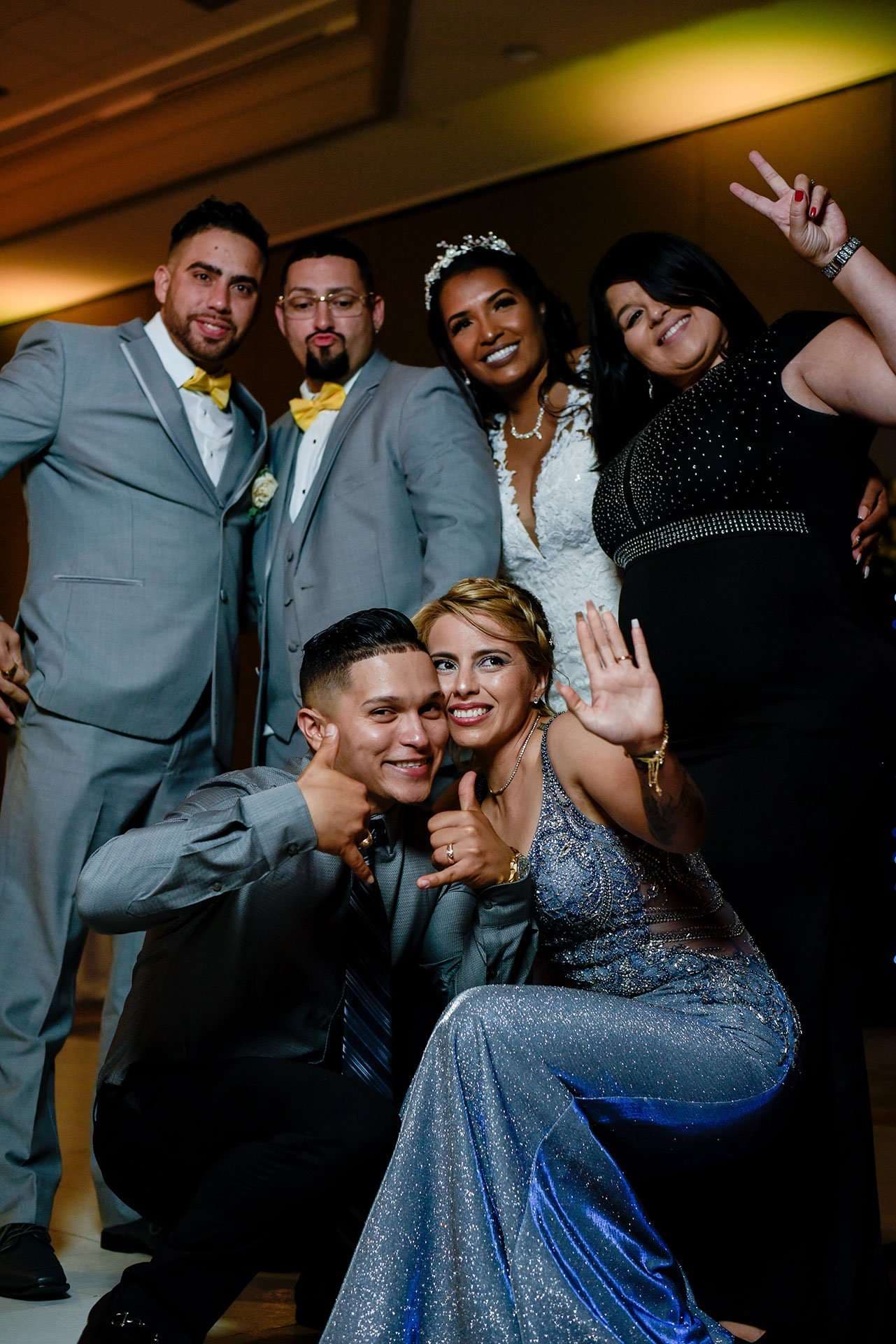 Five wedding guests in formal wear posing playfully together, two men in gray suits with yellow bow ties behind
