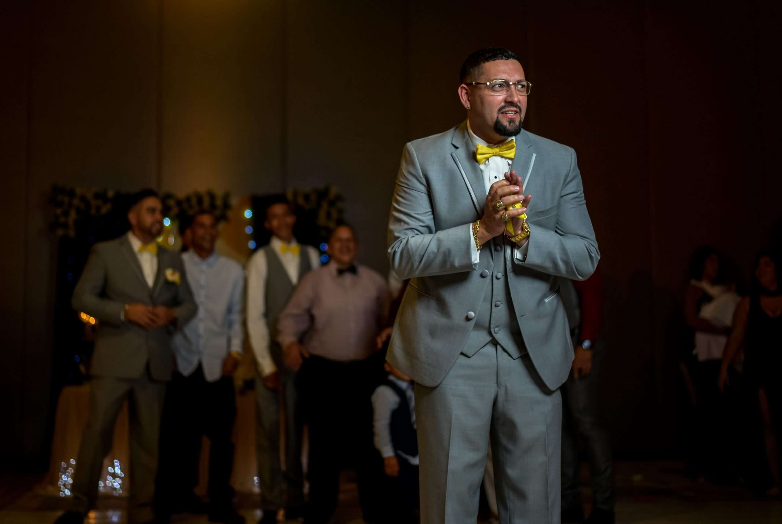 Man in gray suit and yellow bow tie clasping his hands while groomsmen stand smiling behind him in a ballroom
