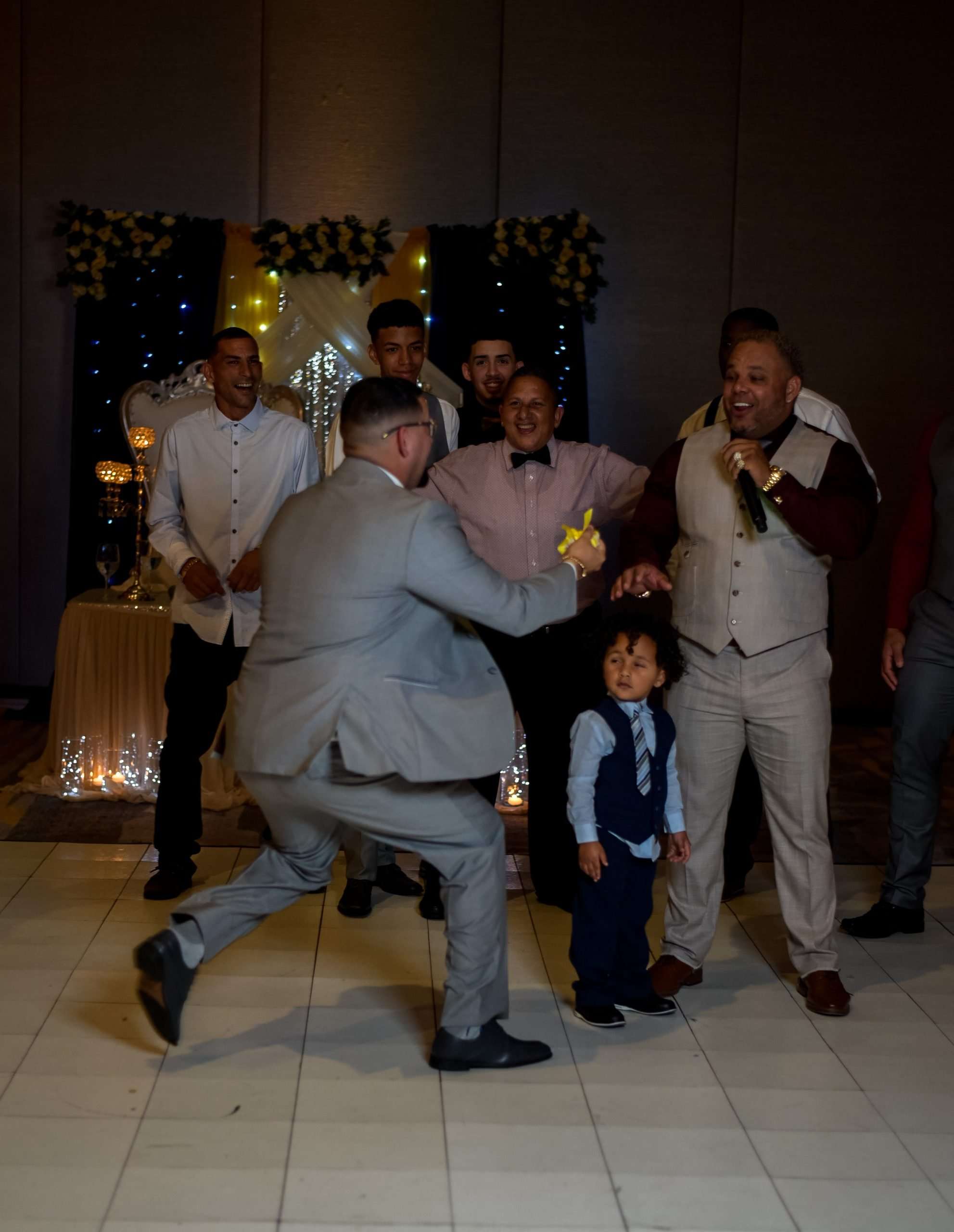 Man in gray suit lunges playfully toward a toddler in a vest as smiling groomsmen watch at a reception
