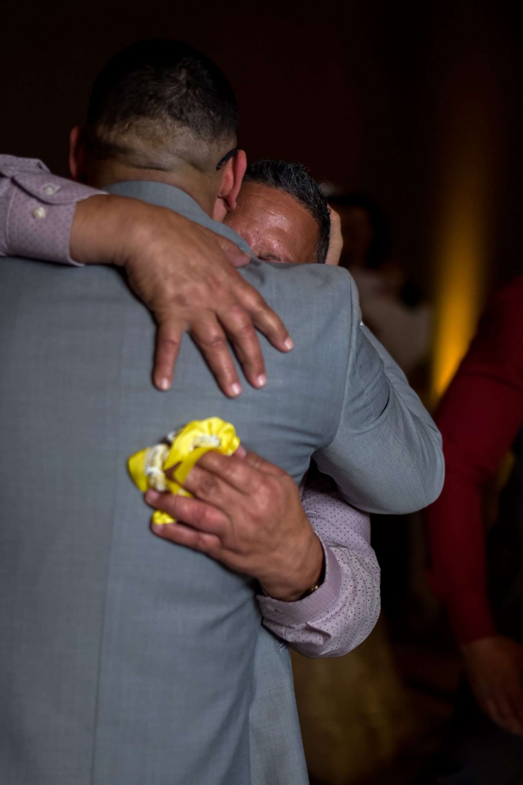 Two men in an emotional embrace at a reception, one in a gray suit holding a yellow handkerchief