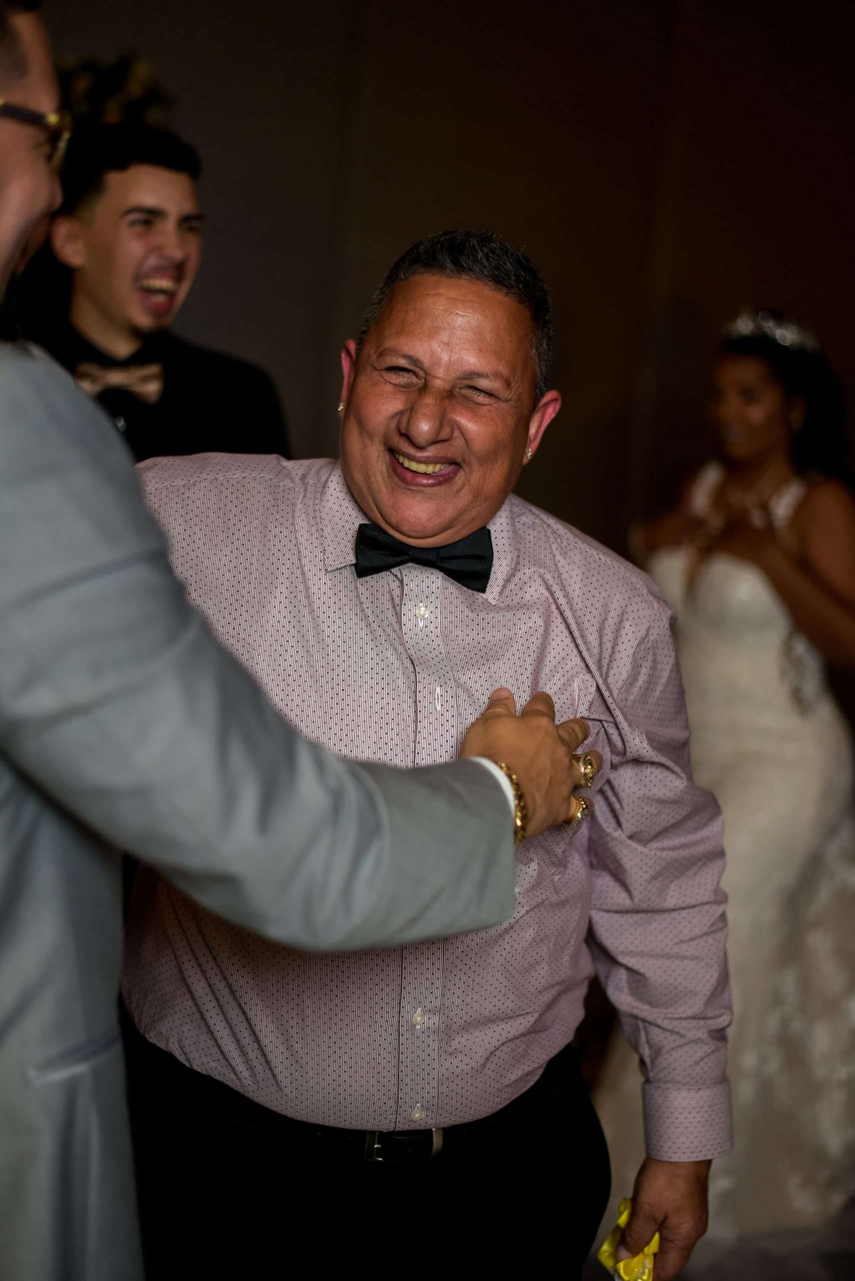Man in a pink dotted shirt and black bow tie laughing joyfully at a wedding reception while guests smile behind him
