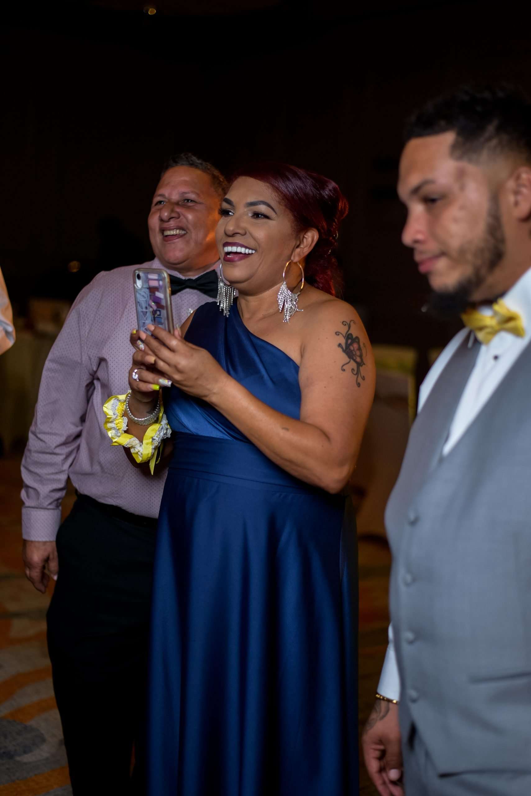 Smiling woman in a royal blue gown films on her phone while two men in bow ties stand beside her at a reception