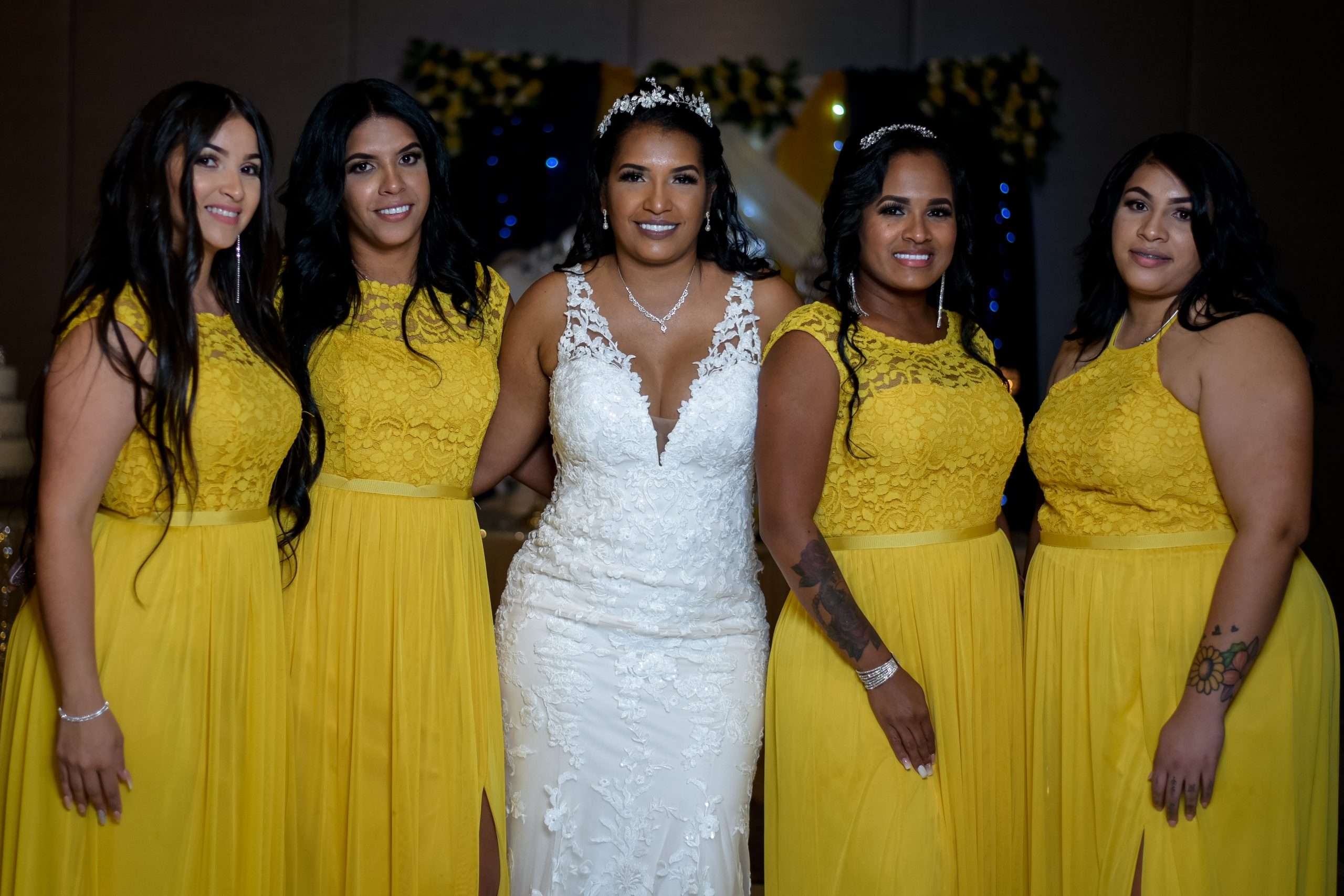Bride in a white lace gown and tiara posing with four bridesmaids in yellow lace dresses at a reception