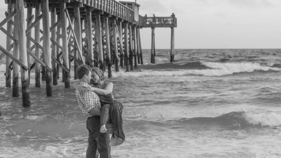 Black and white image of a man lifting his partner in the surf beside a wooden pier
