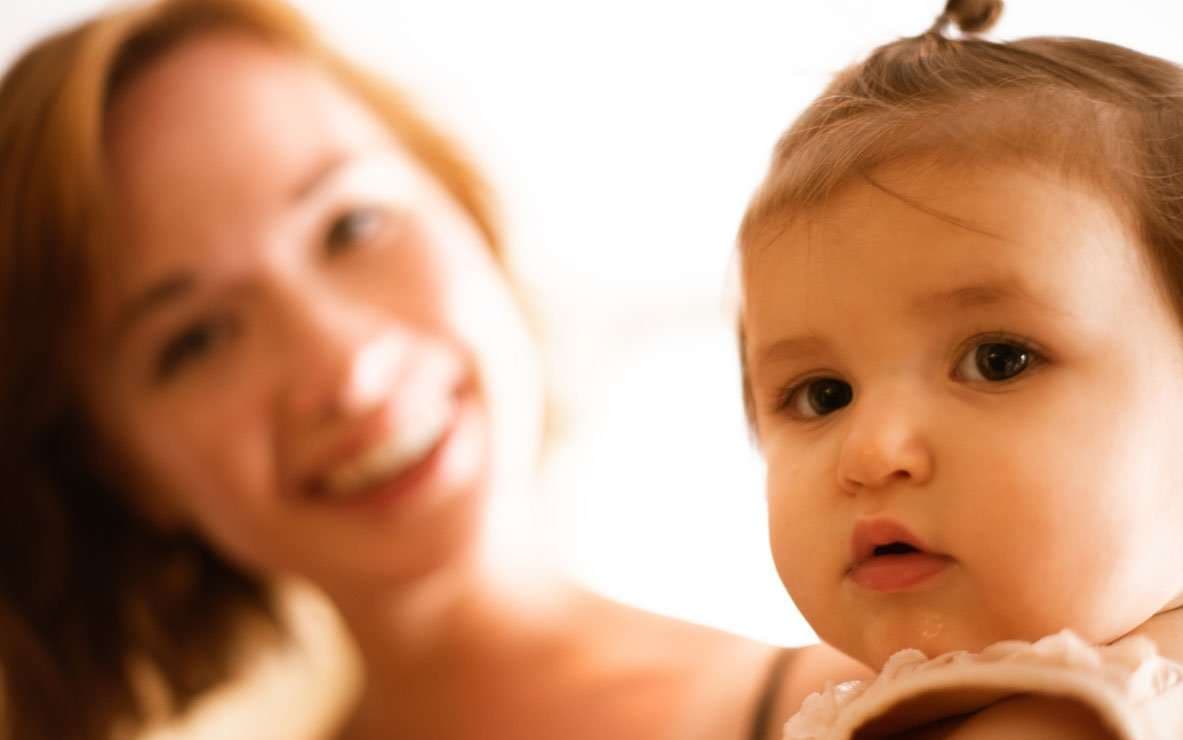Baby with a small top ponytail looking at the camera while a smiling woman is softly blurred behind