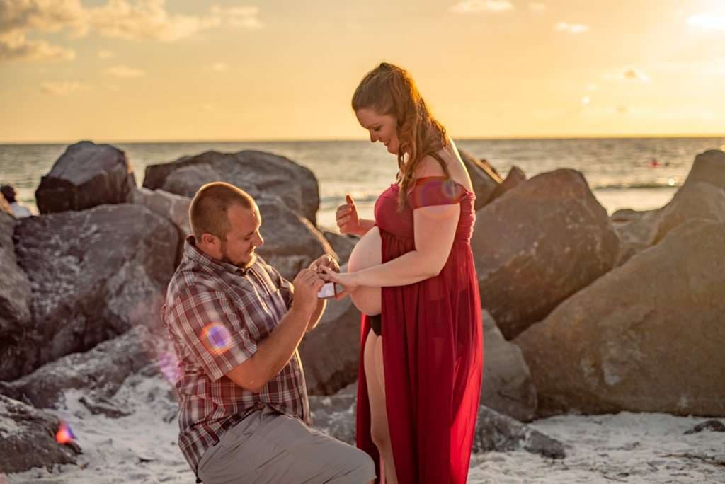 Man kneeling in the sand to propose to his pregnant partner in a red gown, rocks and sunset sea behind them